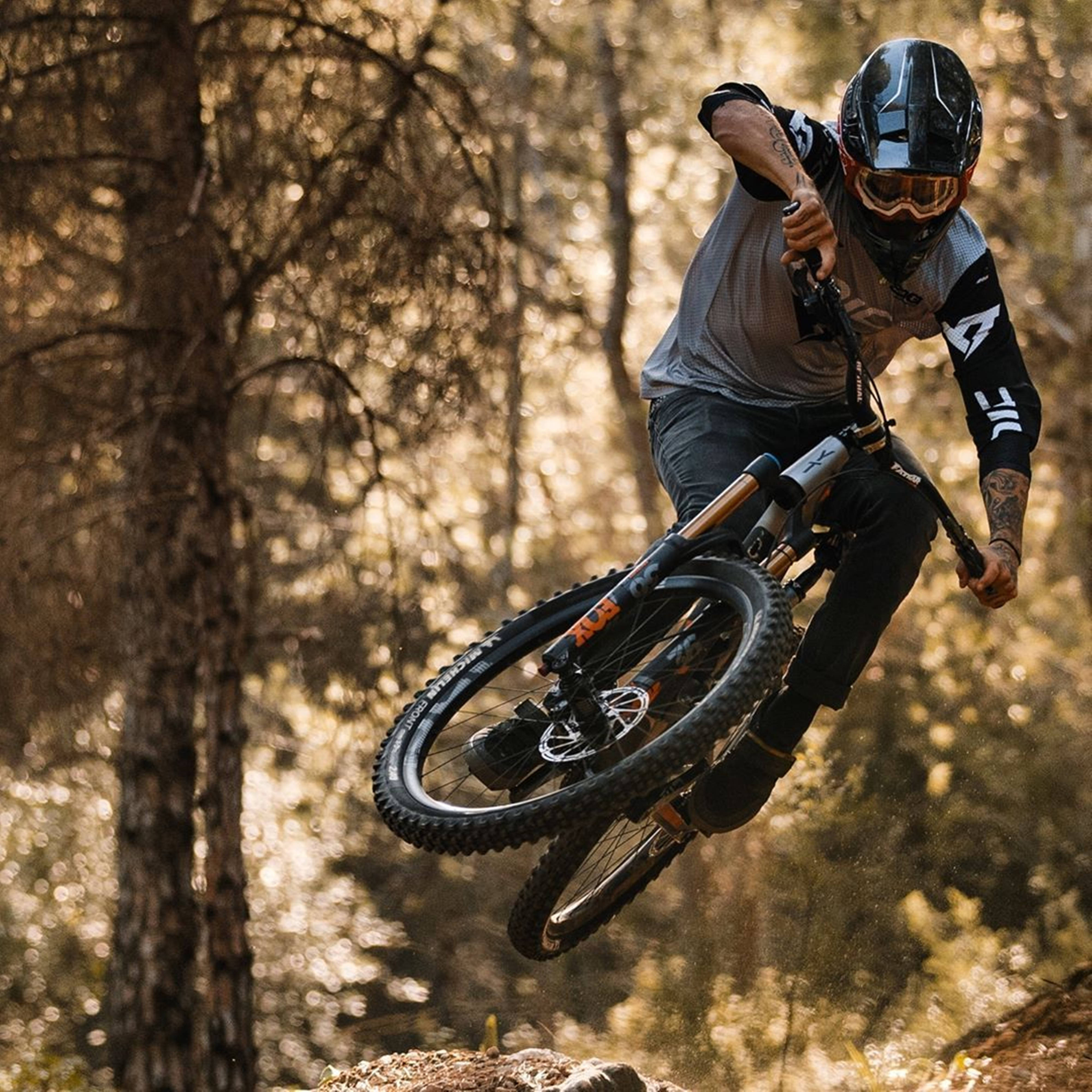 Mountain Bike 桌布 壁紙 — 一名登山車手在森林小徑上空飛翔，金色的陽光穿過樹木灑落。