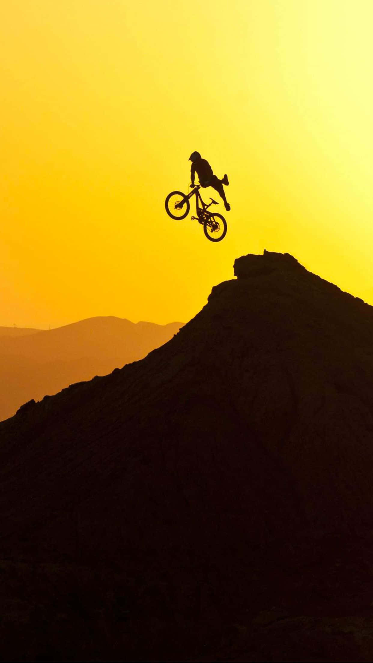 日落 桌布 壁紙 — 一名登山車手在金色夕陽的映襯下， silhouetted 在岩石山峰上空飛翔。