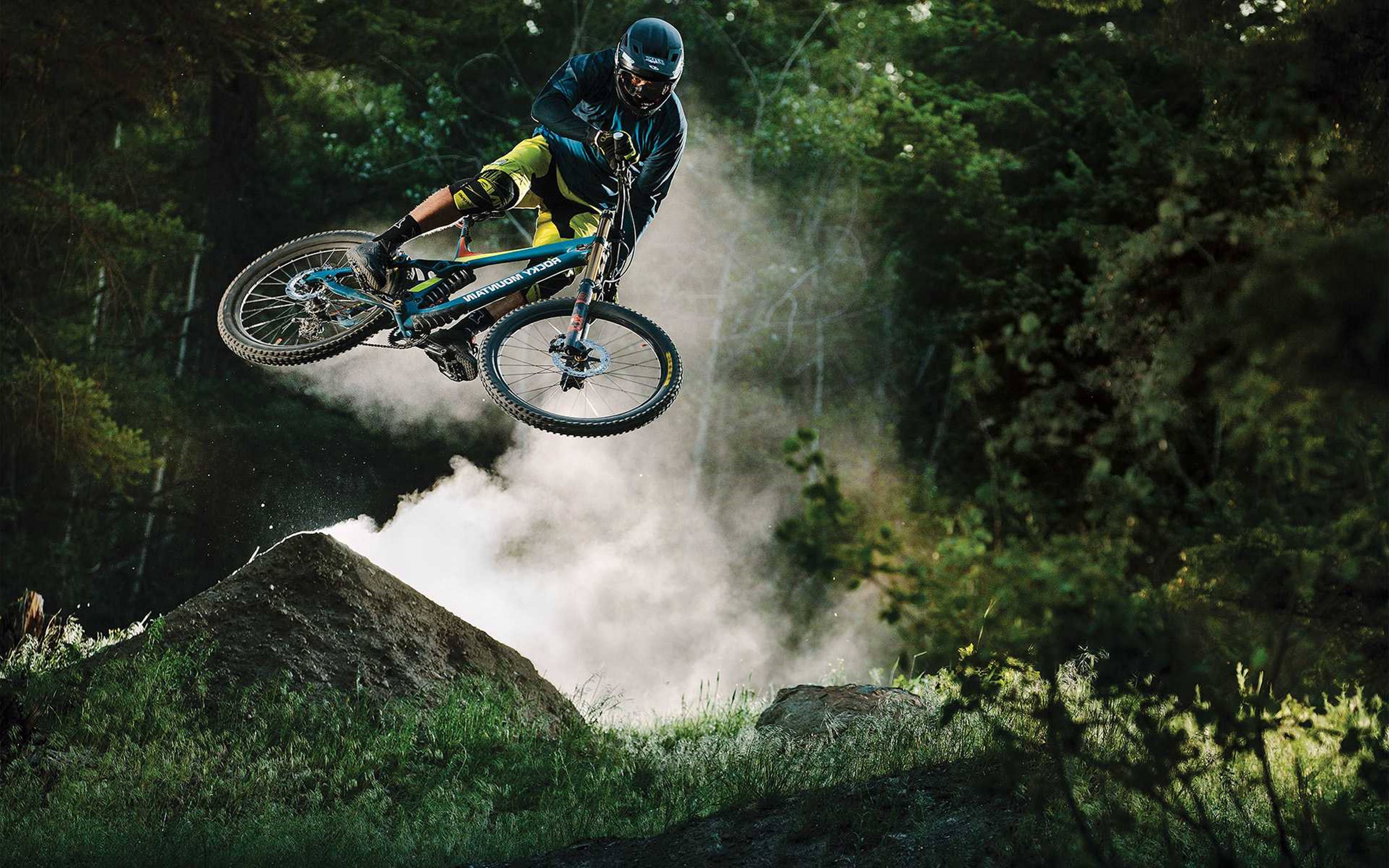 Mountain Bike 桌布 壁紙 — 一名登山車手在陽光普照的森林中飛越土坡跳台，下方塵土飛揚。