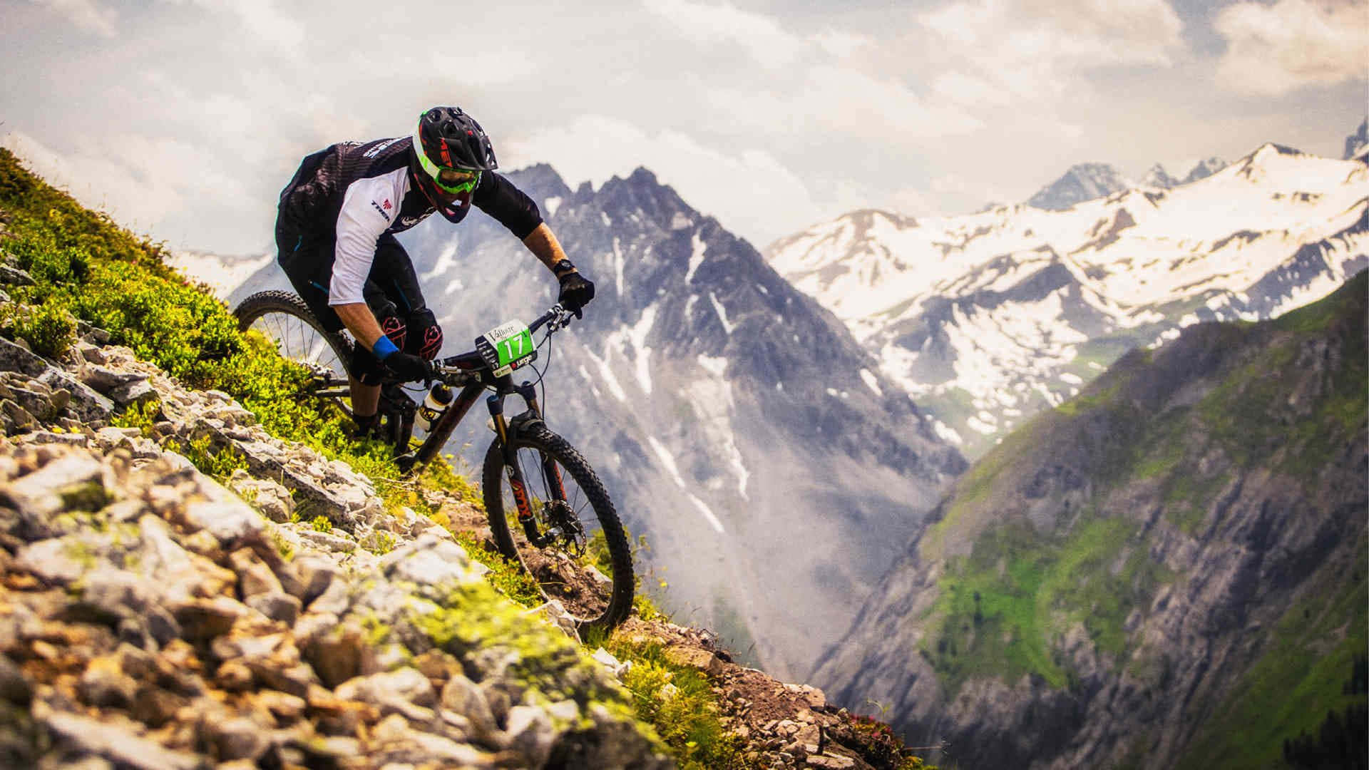Bikes 桌布 壁紙 — 一名登山車騎士在岩石小徑上行駛，背景是白雪覆蓋的山峰和綠色山坡。