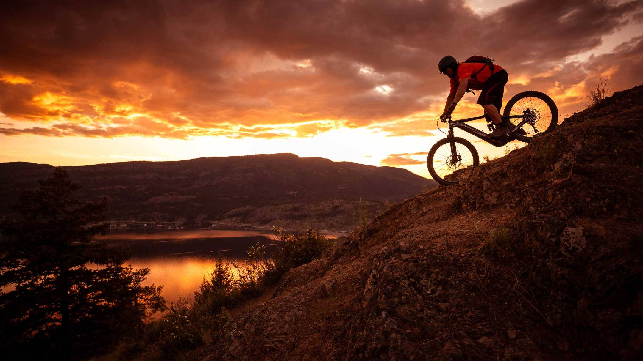 Mountain Bike 桌布 壁紙 — 一名登山車騎士在日落時分騎行在陡峭的岩石小徑上，天空中有火紅的橙色雲彩，下方是倒映著景色的湖泊。