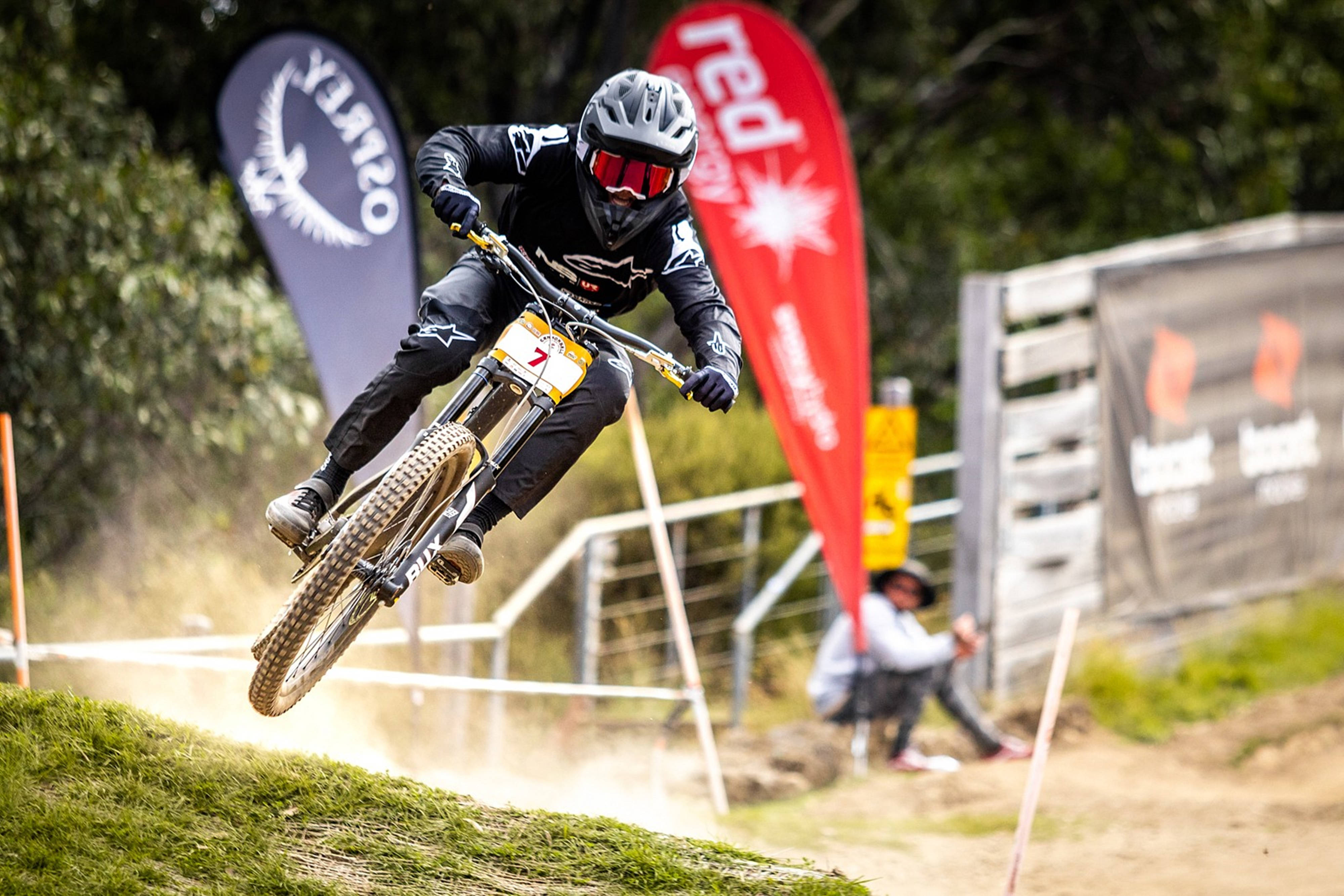 Mountain Bike 桌布 壁紙 — 一名登山車騎士在競爭激烈的下坡賽中，於草地小徑上空飛翔，背景有贊助商橫幅。