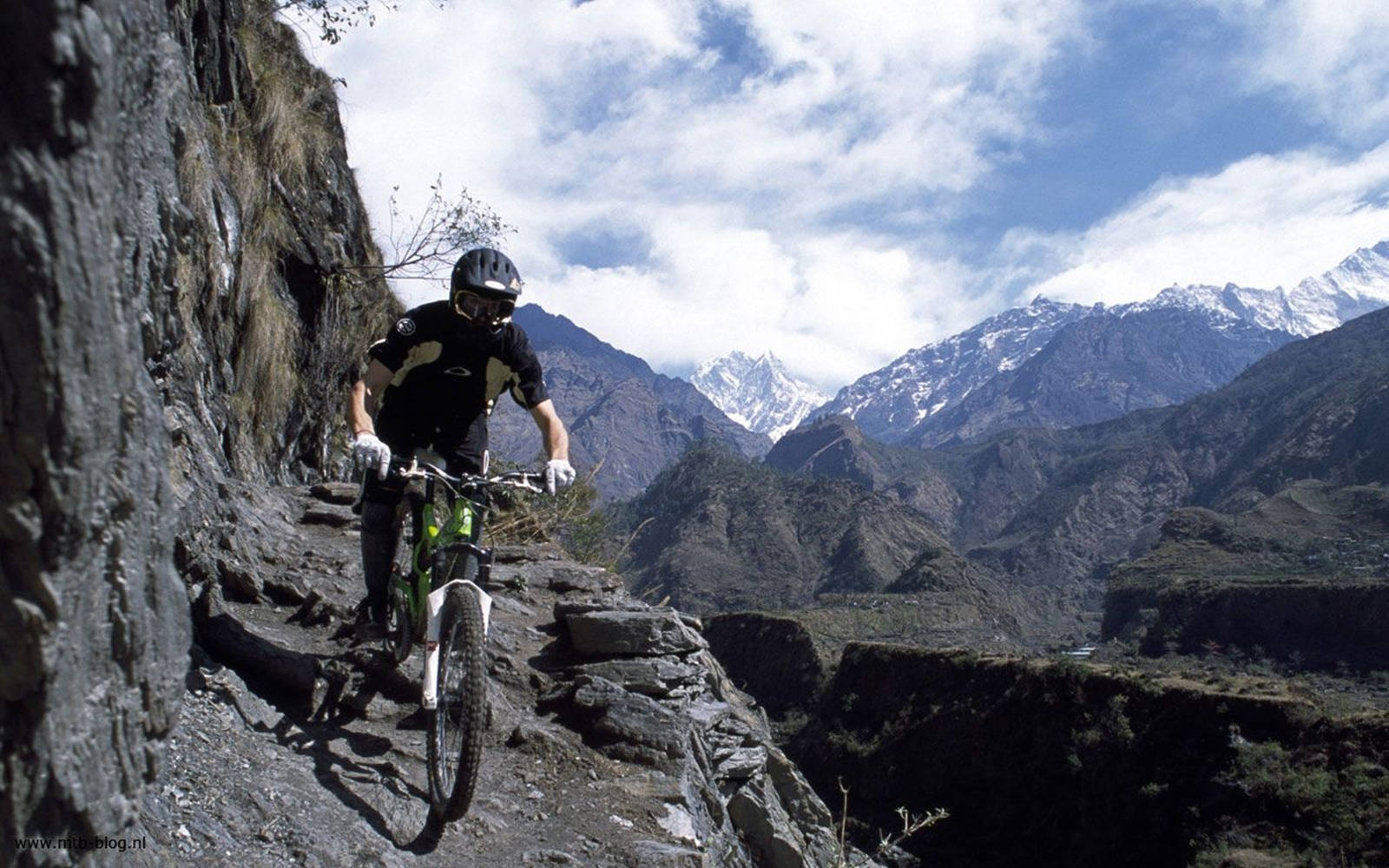 Mtb 桌布 壁紙 — 一名登山車騎士在部分多雲的天空下，沿著陡峭懸崖旁的狹窄岩石小徑行駛，背景中聳立著白雪覆蓋的山峰。