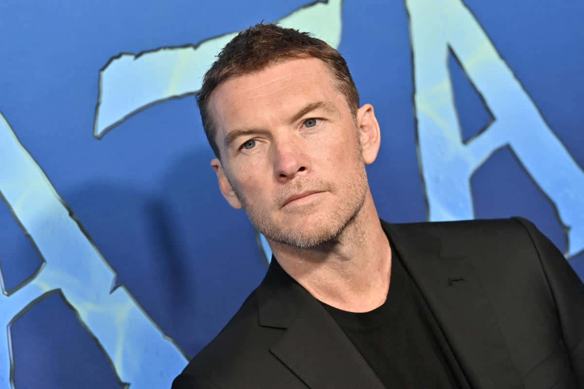 actor 桌布 壁紙 — 一名短棕髮、鬍渣的男子，身穿黑色西裝外套配黑色襯衫，背景為藍色，帶有大型風格化字母。