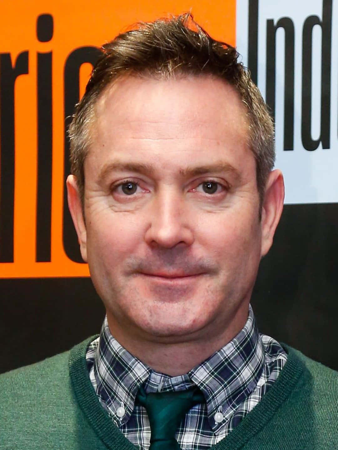thomas lennon 桌布 壁紙 — 一名短棕髮男子穿著綠色毛衣，內搭格子襯衫和綠色領帶，背景為橙色和白色。
