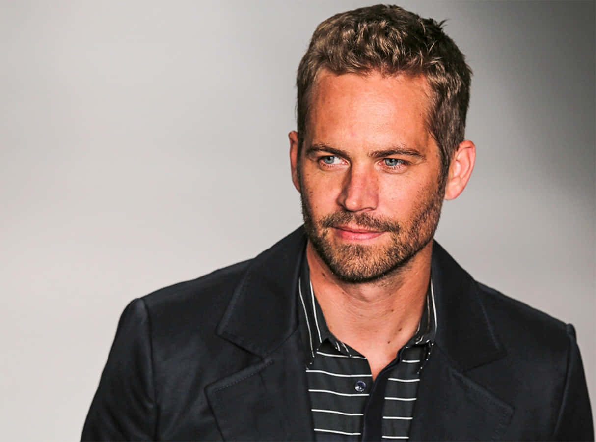 Paul Walker 桌布 壁紙 — 一名短棕髮留鬍鬚的男子，在純灰色背景前穿著深色西裝外套配條紋襯衫。