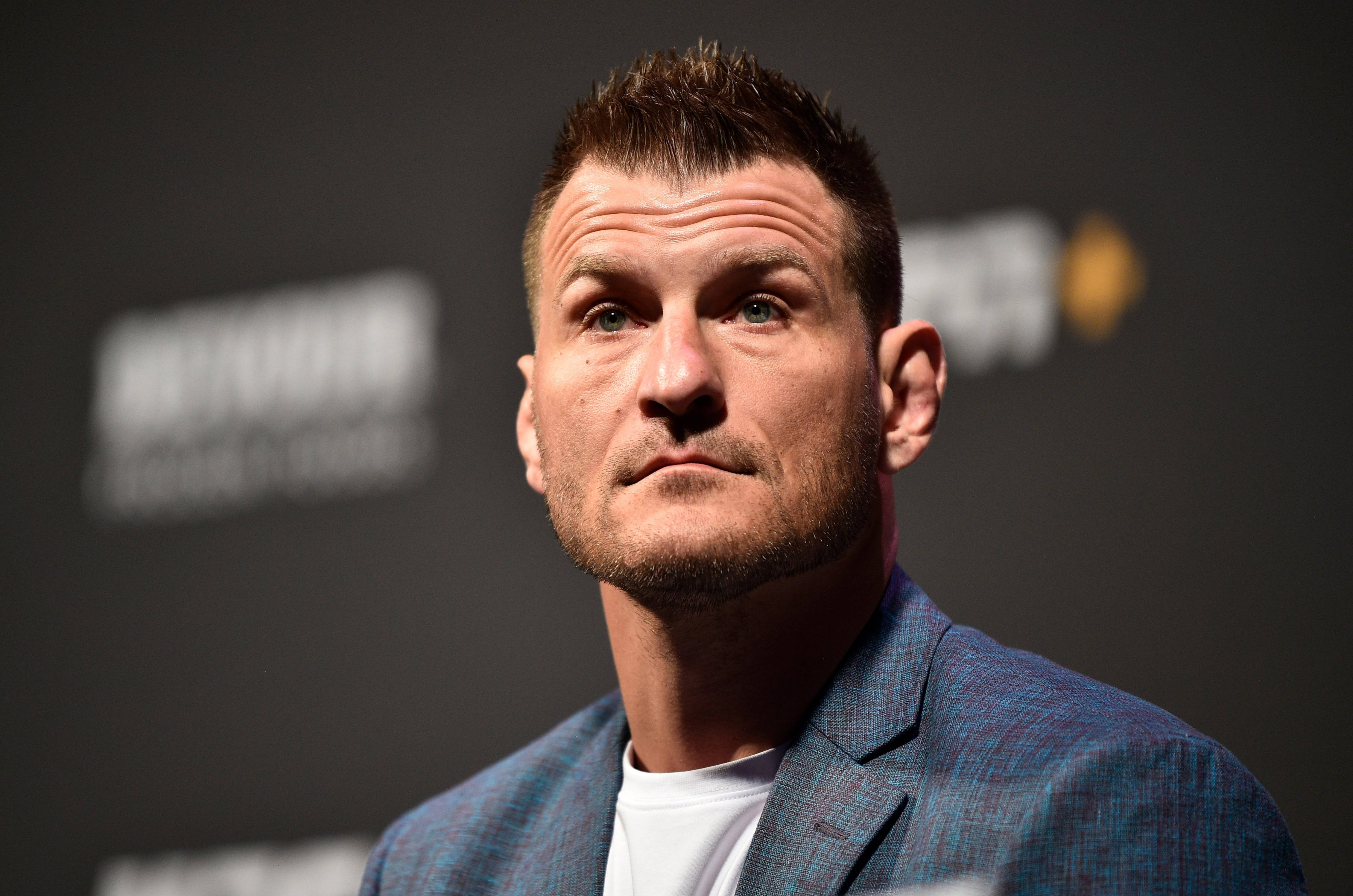 UFC 桌布 壁紙 — 一名短髮尖刺、藍眼深邃的男士特寫肖像，身穿藍色紋理西裝外套內搭白色T恤，背景為深色帶模糊標誌。
