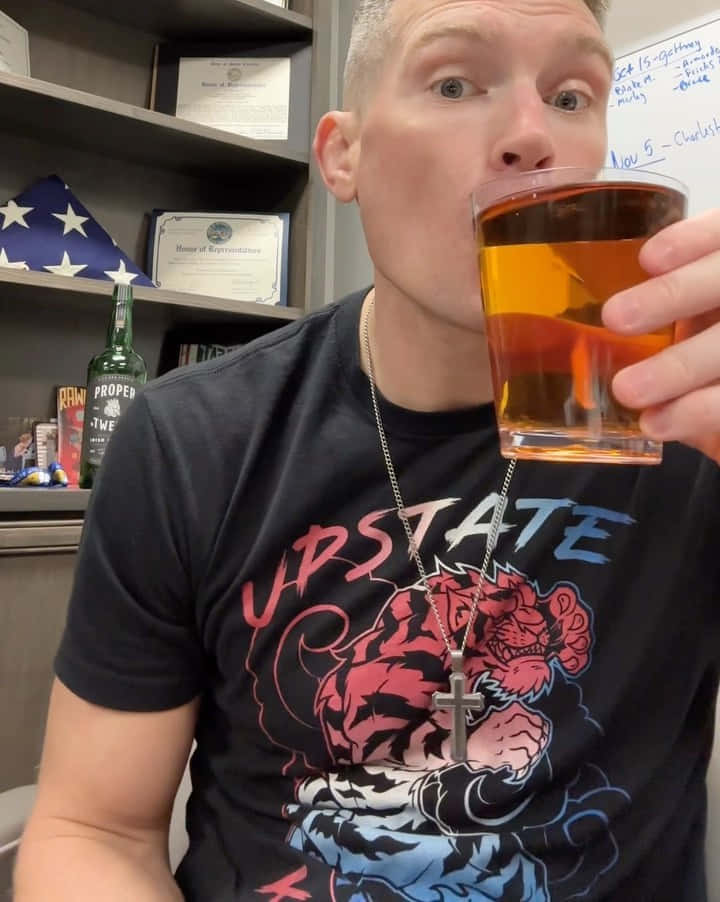 Beer 桌布 壁紙 — 一名短髮男子在室內穿著圖案T恤和十字架項鍊，從玻璃杯啜飲琥珀色液體。