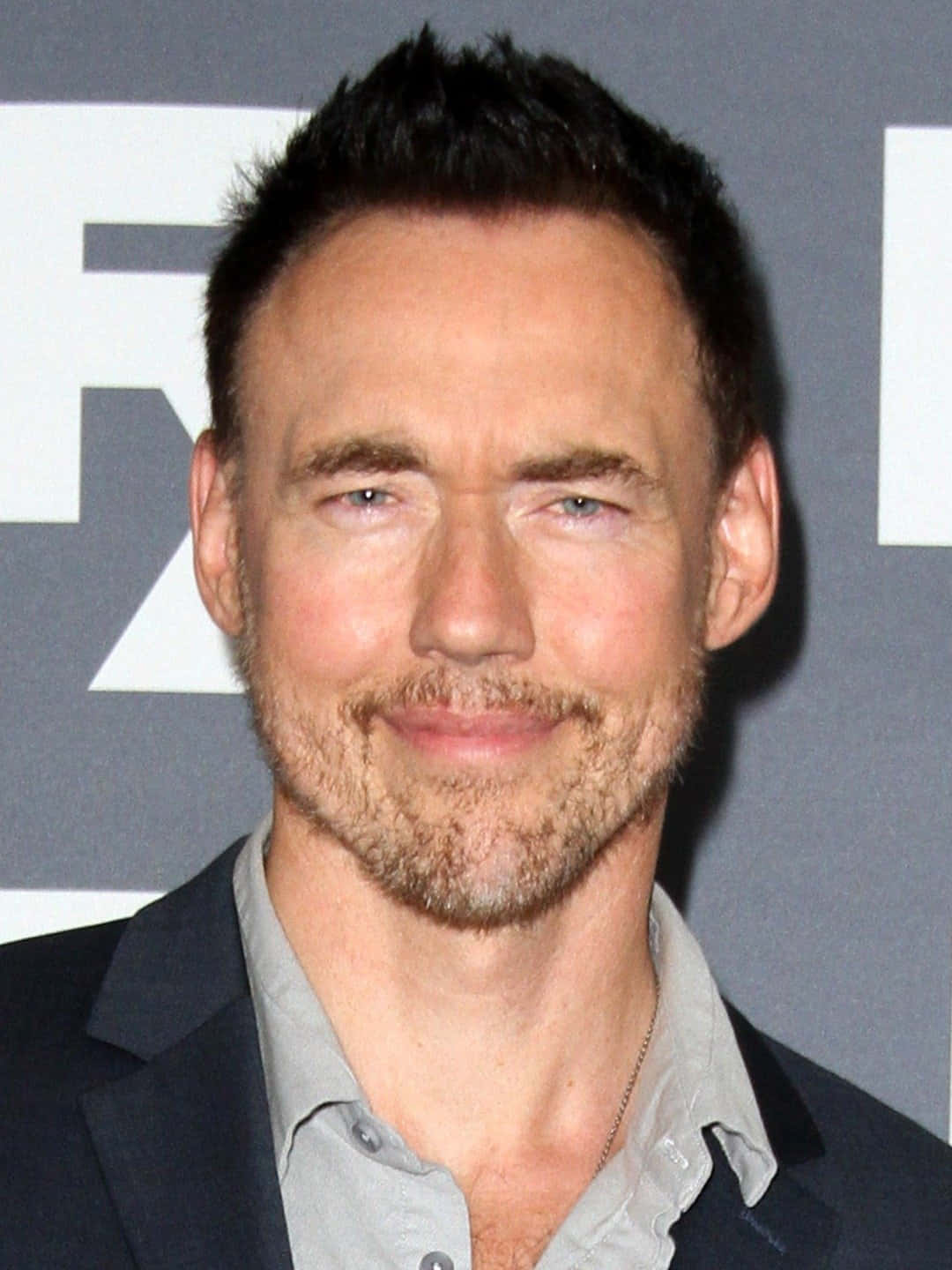 Kevin Durand 桌布 壁紙 — 一名短黑髮、留著淺鬍鬚的男士，對著鏡頭溫暖微笑的特寫肖像。