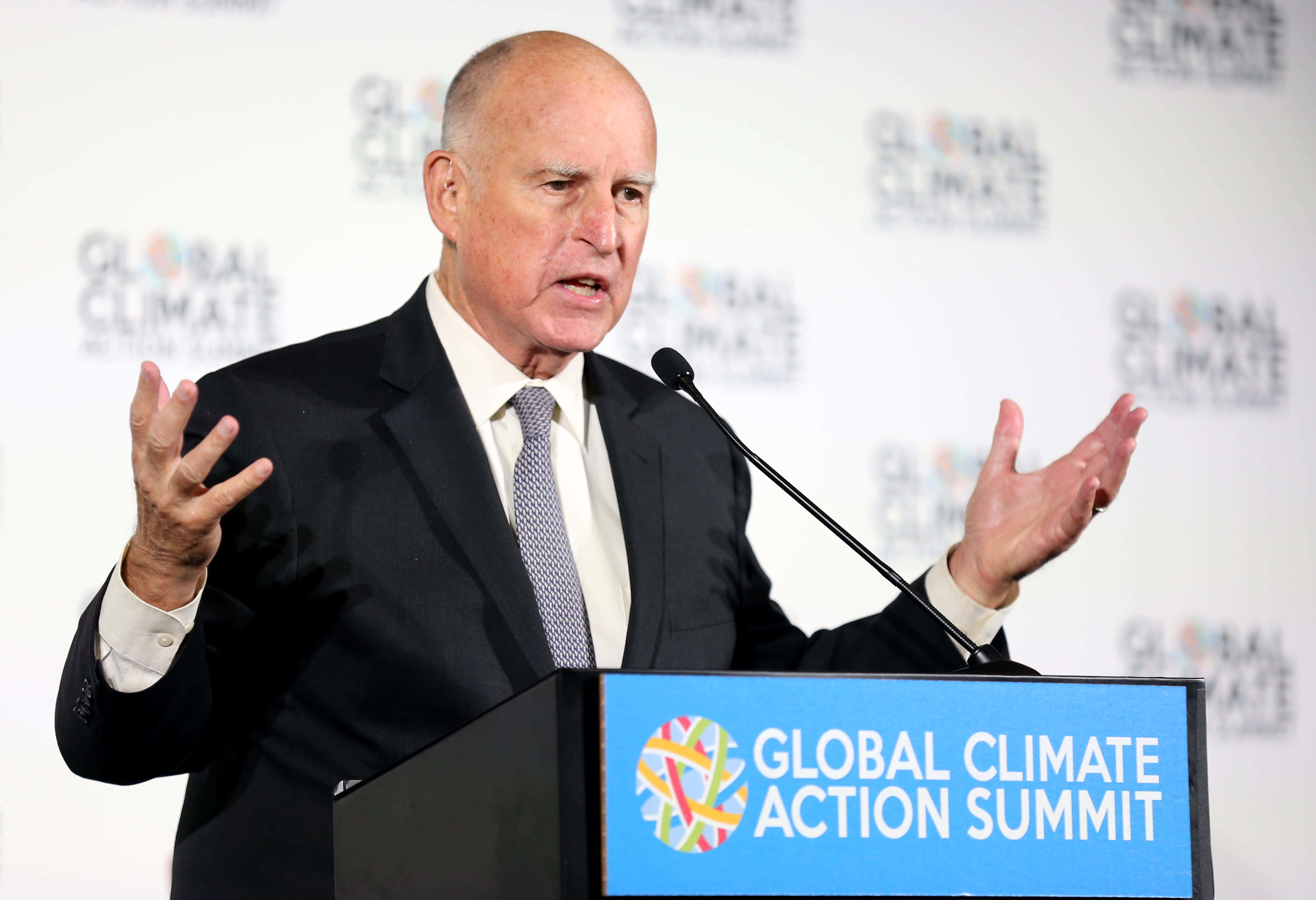 政治 桌布 壁紙 — 一名禿頭男子身穿深色西裝，在標有 Global Climate Action Summit 的講台上熱情地做手勢發言。