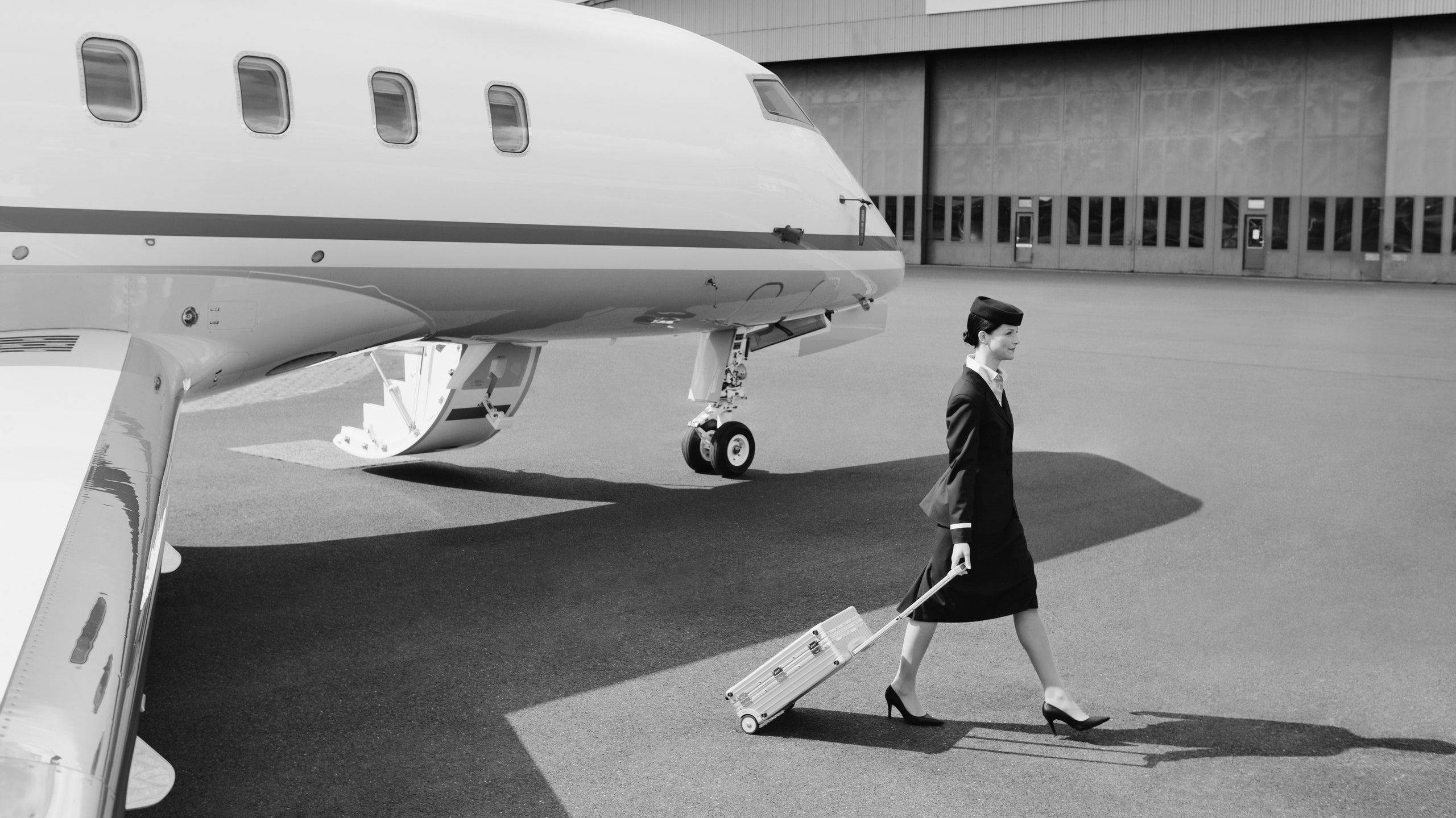飛機 桌布 壁紙 — 一名空姐自信地拖著滾輪行李箱，在機場停機坪上的一架私人飛機旁行走，黑白照片。