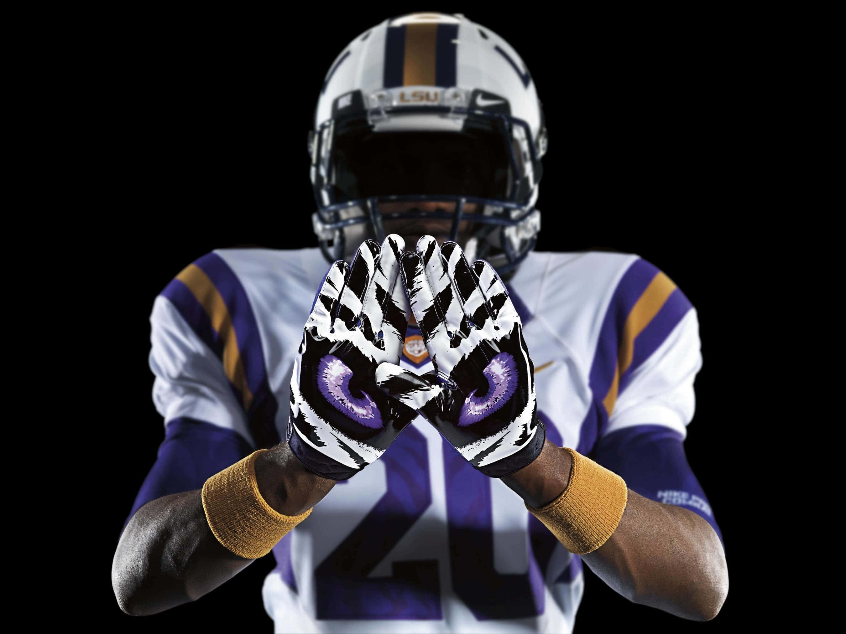 Lsu Tigers 桌布 壁紙 — 一名穿著LSU裝備的足球運動員在黑色背景前舉起帶有紫色眼睛圖案的手。