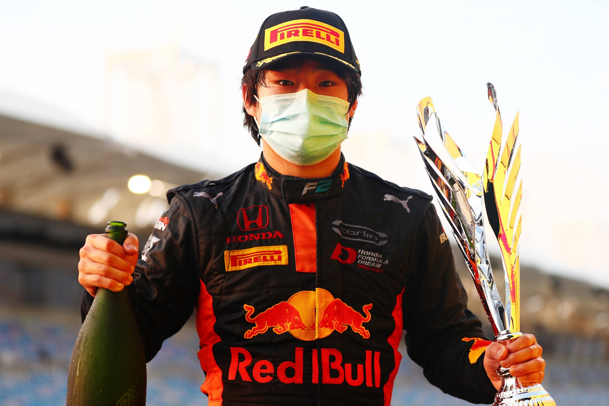 Red Bull 桌布 壁紙 — 一名穿著Red Bull賽車服的二級方程式車手在賽道上手持獎盃和香檳瓶，同時戴著口罩。