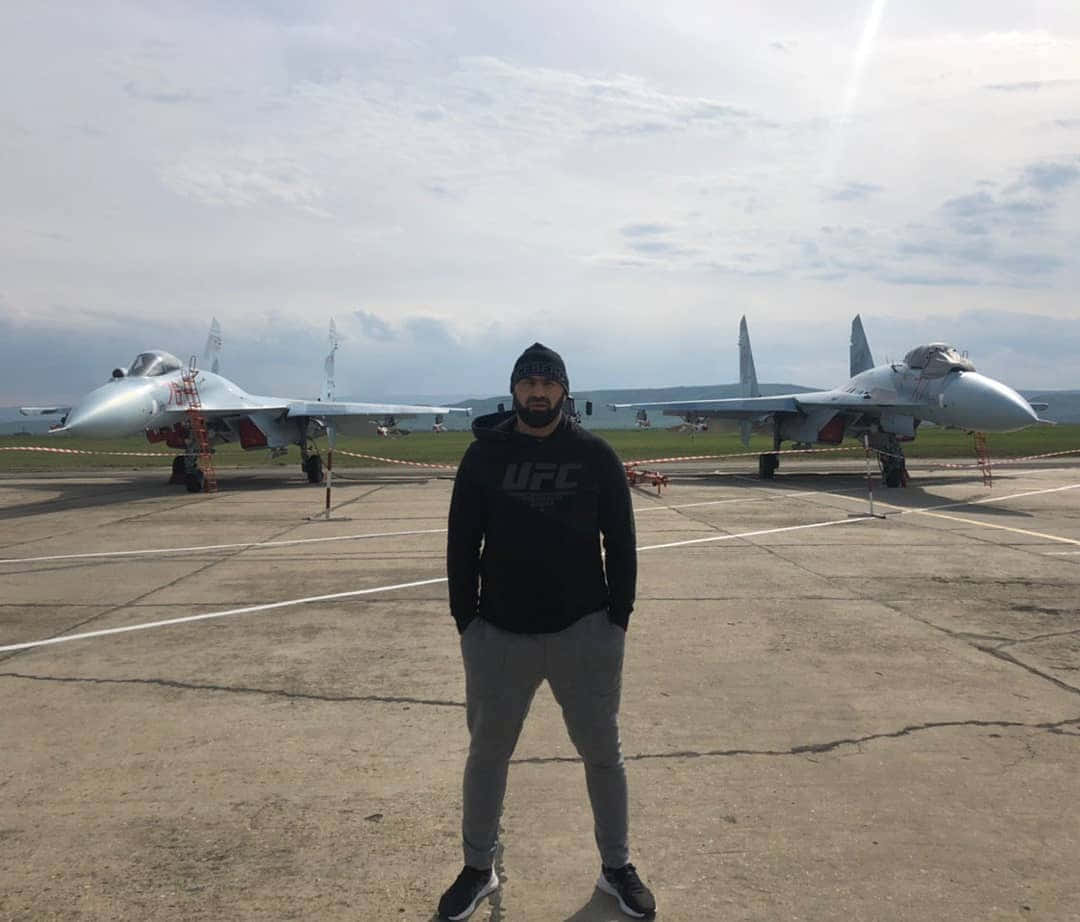 MMA 桌布 壁紙 — 一名穿著UFC連帽衫的男子自信地站在機場上，身後停著兩架戰鬥機，天空多雲。