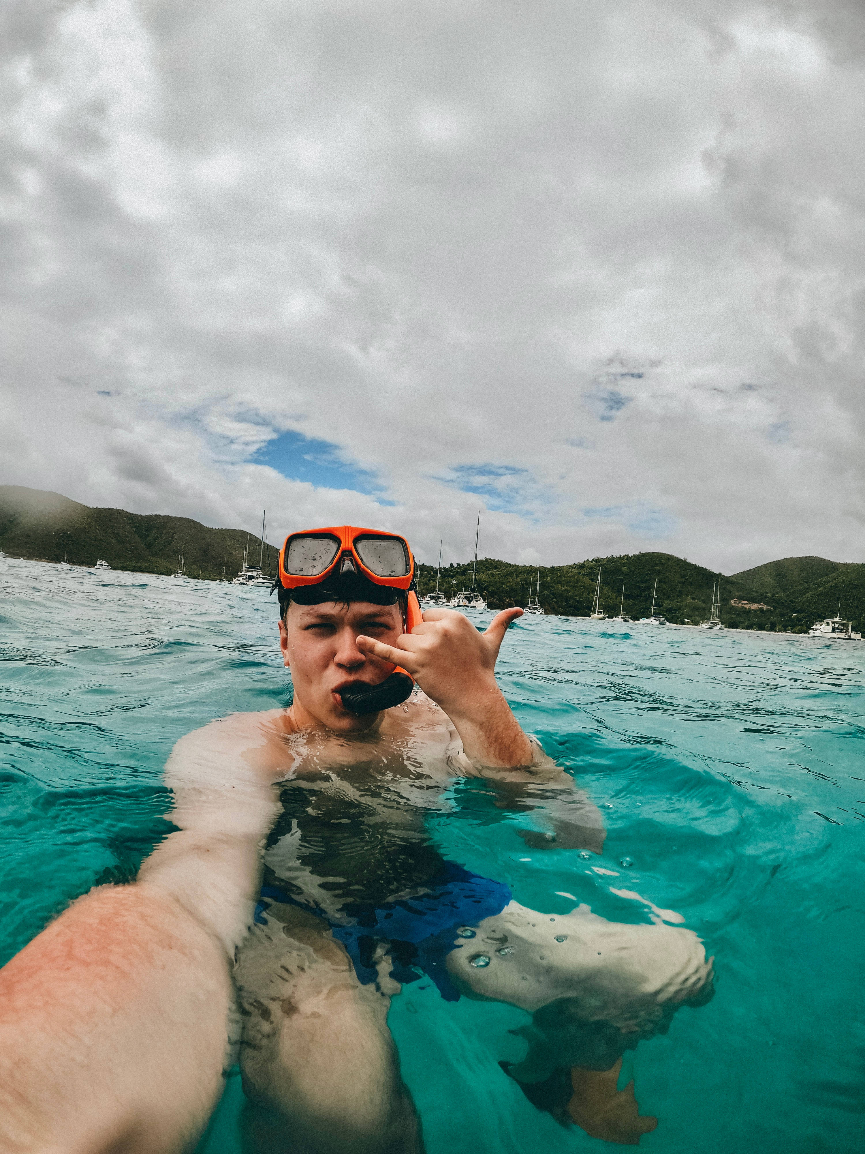 Snorkeling 桌布 壁紙 — 一名穿著亮橙色浮潛裝備的人微笑著比出和平手勢，漂浮在碧綠的海水中，背景有帆船和綠色山丘。