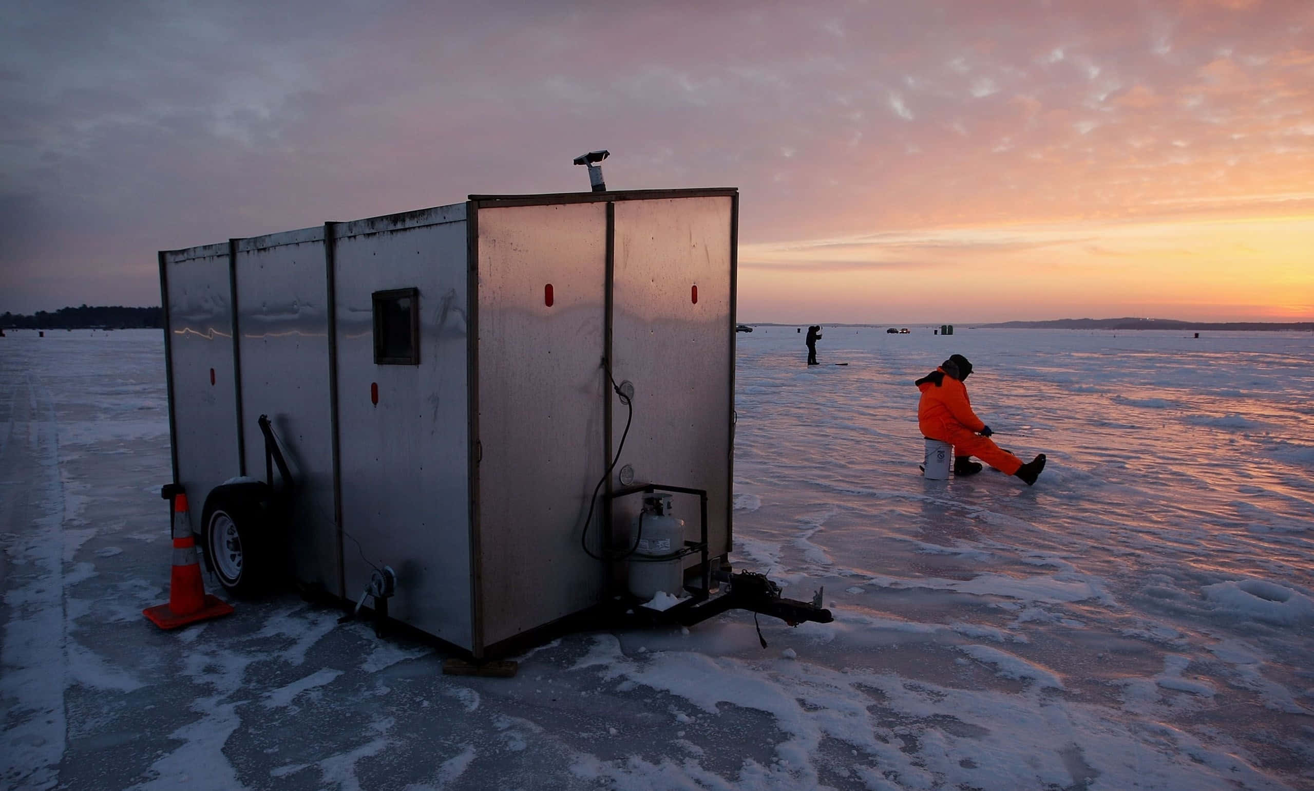 Ice Fishing 桌布 壁紙 — 一名穿著亮橙色裝備的人坐在冰上，旁邊有一個金屬拖車，熾熱的夕陽為冰凍的湖面和天空塗上色彩。