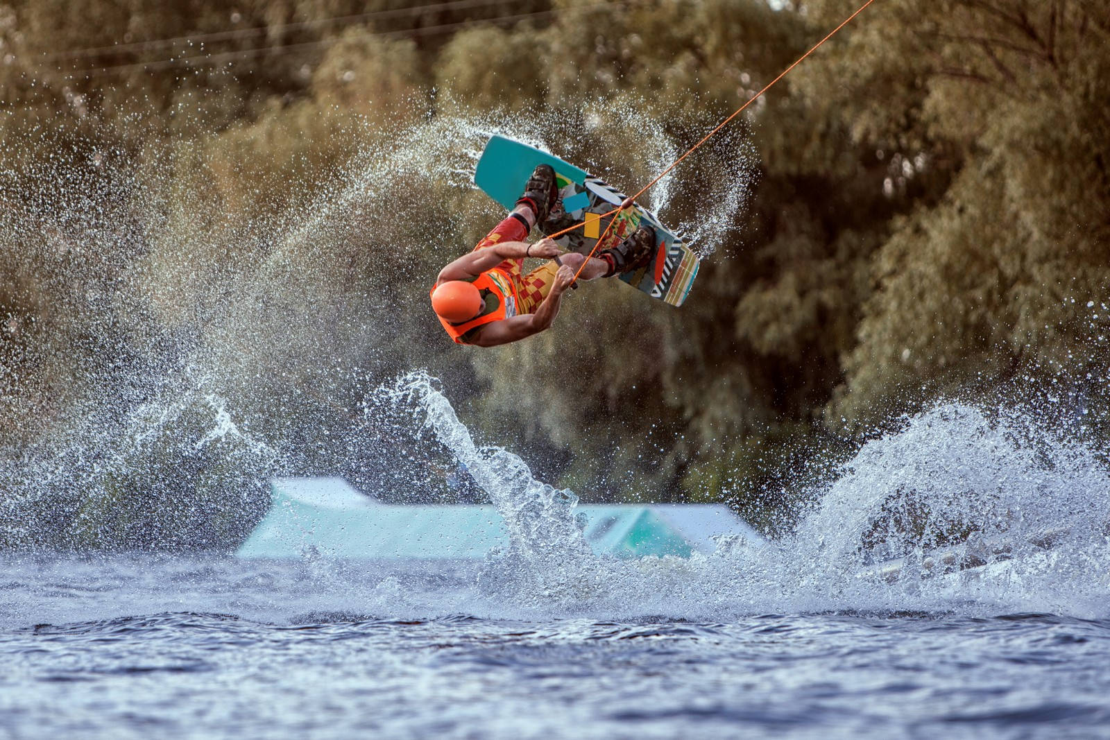 surfing 桌布 壁紙 — 一名穿著亮橙色裝備的滑水板運動員在波濤洶湧的藍色水面上執行空中翻轉，背景是濺起水花的綠色樹木。