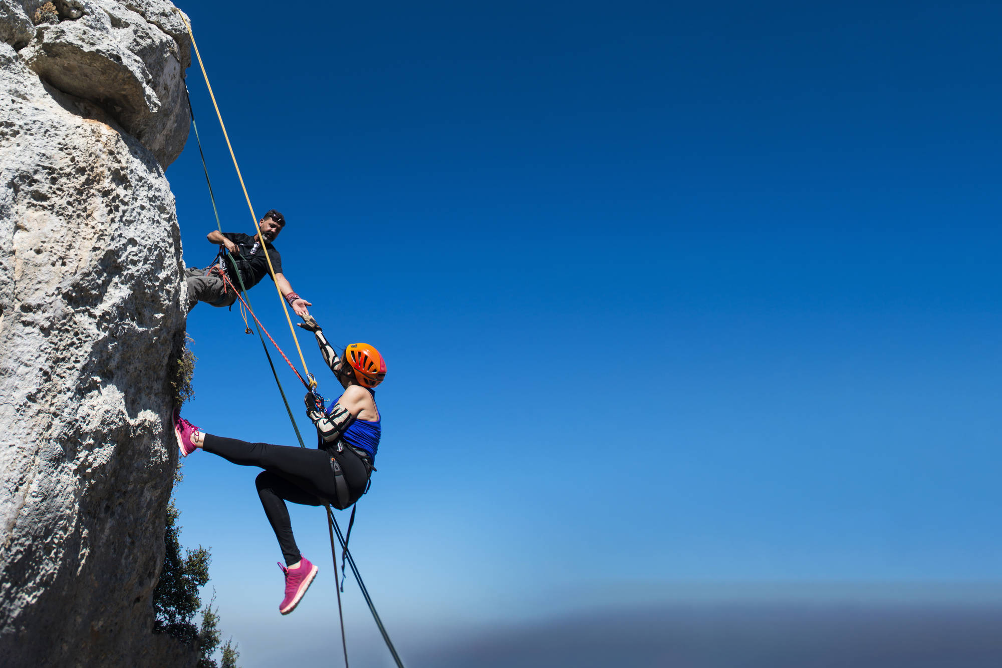 Climbing 桌布 壁紙 — 一名穿著亮粉紅色鞋的攀岩者正在攀登崎嶇的岩壁，而一名確保員在廣闊的晴朗藍天下用繩索保護她。