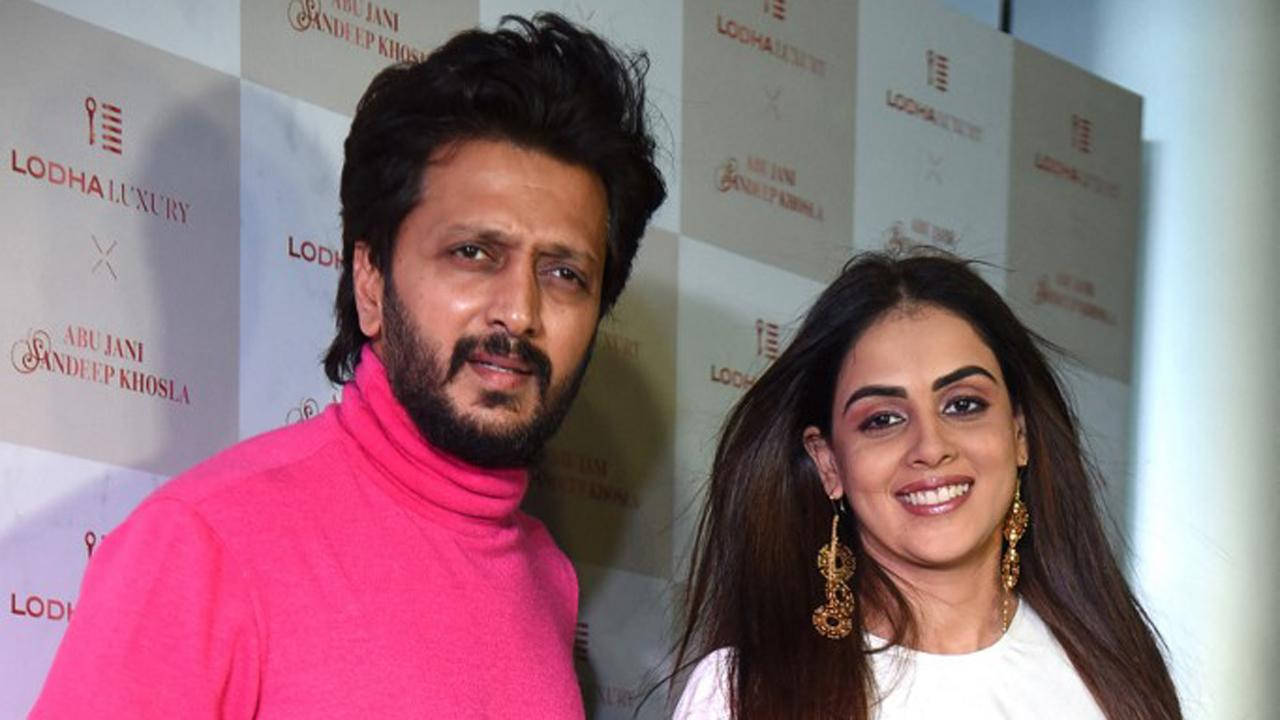 riteish deshmukh 桌布 壁紙 — 一名穿著亮粉紅色高領毛衣的男子和一名穿著白色衣服的女子在品牌活動背景前對著鏡頭微笑。