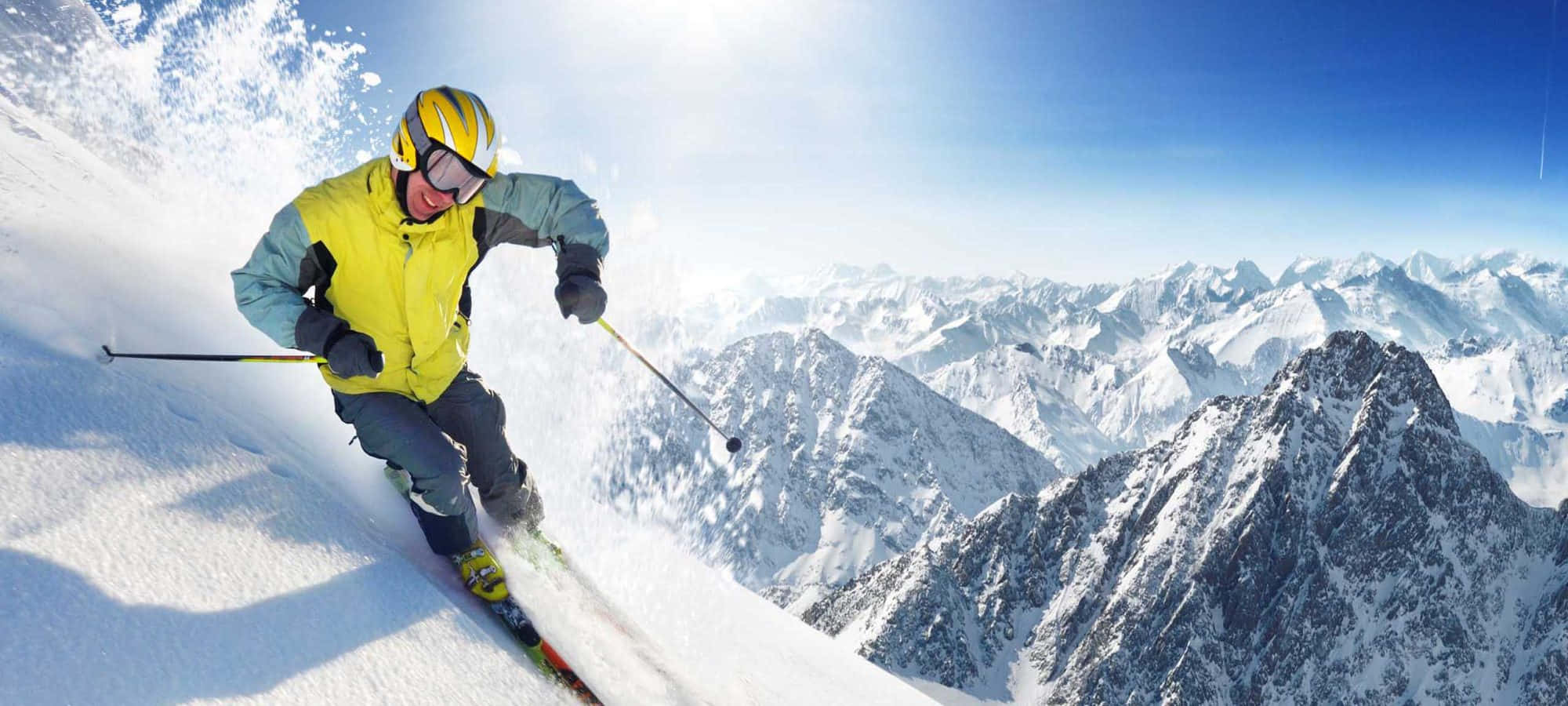 Ski Mountain 桌布 壁紙 — 一名穿著亮黃色外套的滑雪者在晴朗的藍天下，在積雪的山坡上刻滑，雪花飛揚。