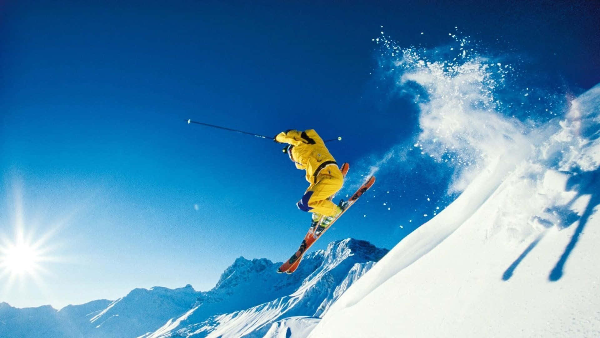 Ski Mountain 桌布 壁紙 — 一名穿著亮黃色滑雪服的滑雪者在晴朗藍天和陽光照耀下，飛越白雪覆蓋的山坡上空。