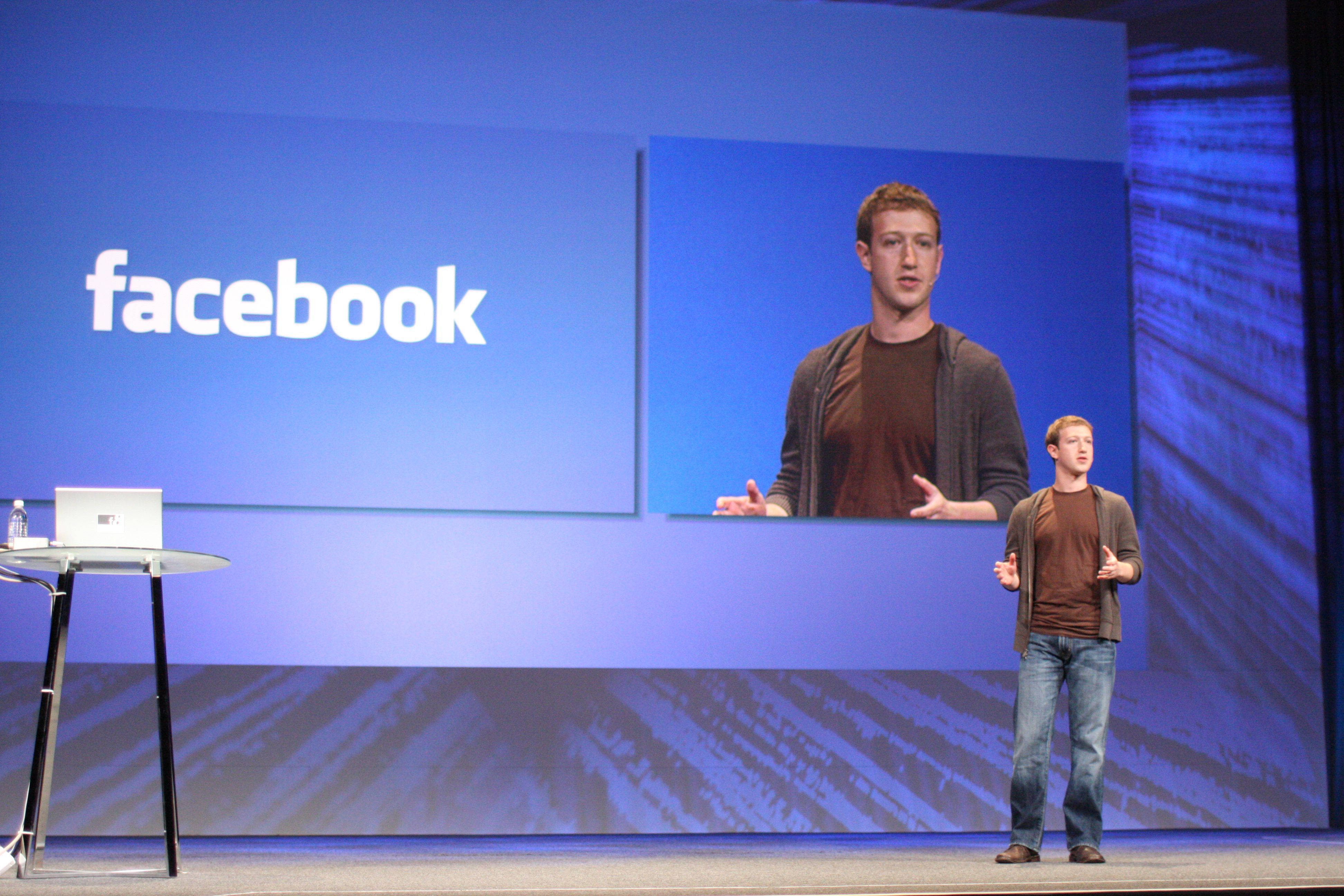 Zuckerberg 桌布 壁紙 — 一名穿著休閒服裝的男子站在舞台上進行簡報，身後的大螢幕顯示Facebook標誌和他的圖像。