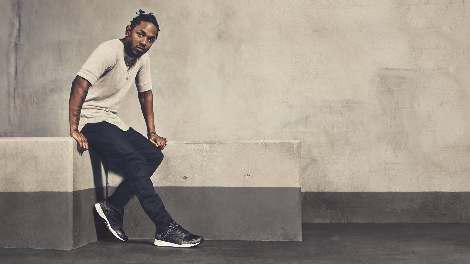 Kendrick 桌布 壁紙 — 一名穿著休閒服裝的男子靠在混凝土長椅上，倚著一面有紋理的牆壁，身處簡約的城市環境中。