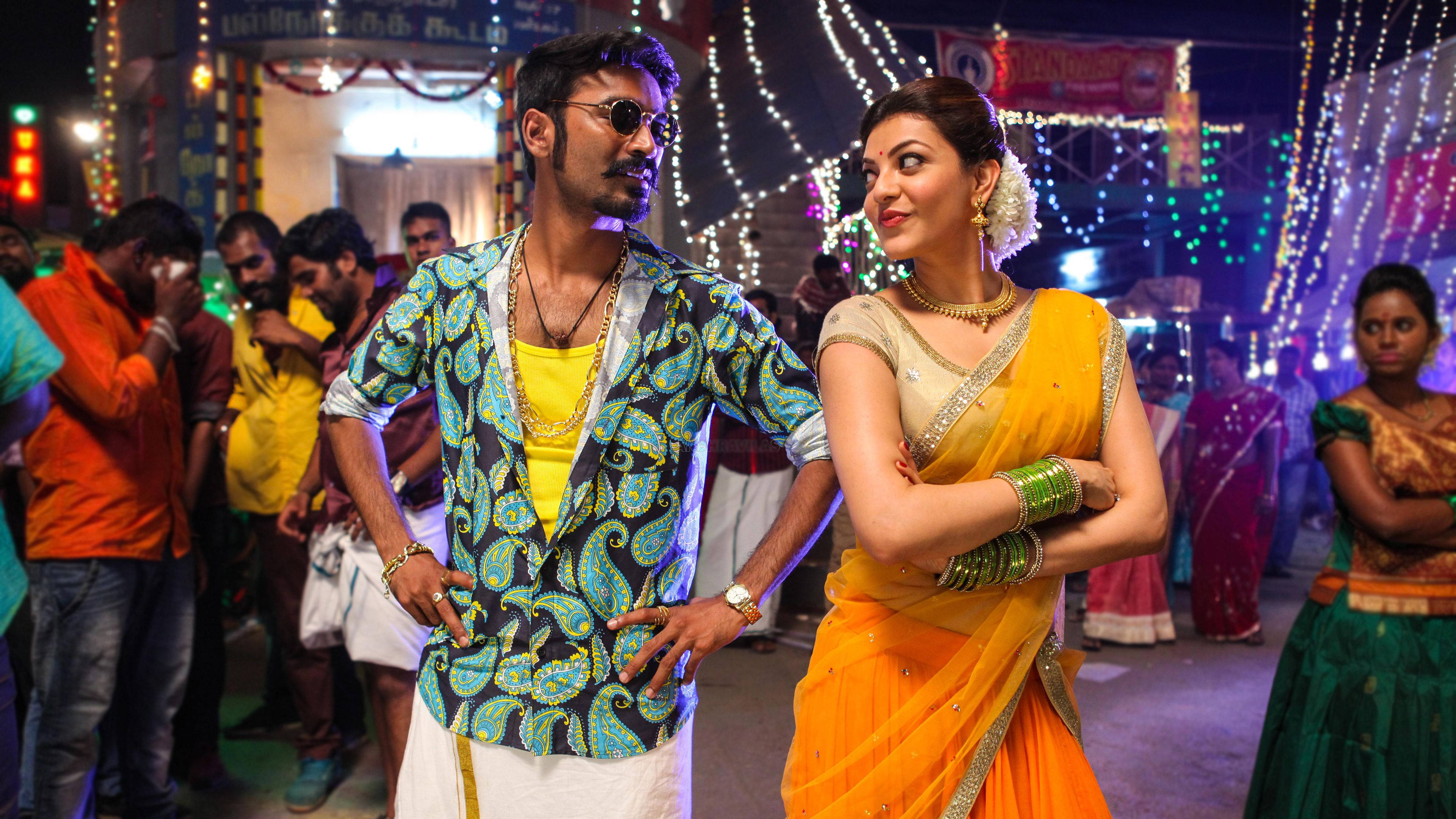 Bollywood HD 桌布 壁紙 — 一名穿著佩斯利夾克的男子和一名穿著黃色紗麗的女子自信地站在一個充滿活力的夜間街頭節日中，周圍有燈串和旁觀者。