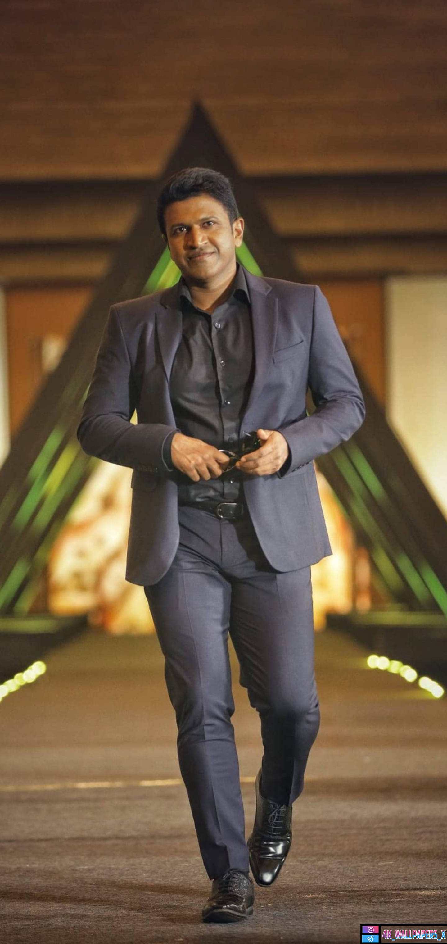 Puneeth Rajkumar 桌布 壁紙 — 一名穿著俐落灰色西裝的男子自信地走在燈光璀璨的舞台上，身後有發光的綠色三角形燈光。