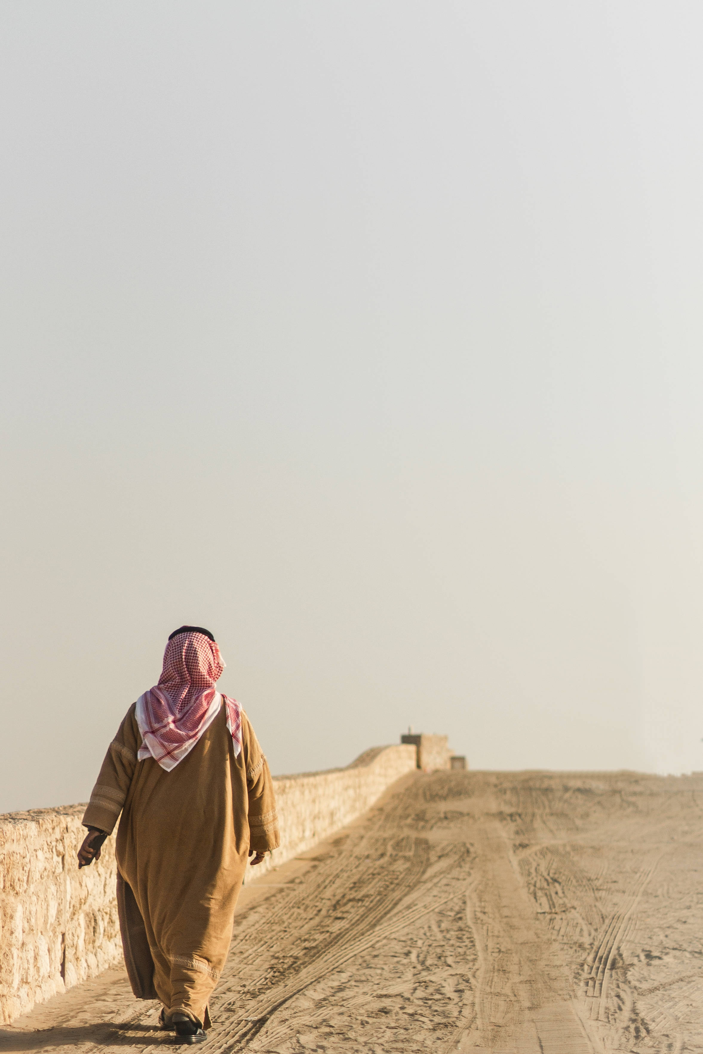 Bahrain 桌布 壁紙 — 一名穿著傳統服飾的男子在朦朧的天空下，獨自沿著塵土飛揚的沙漠牆行走。