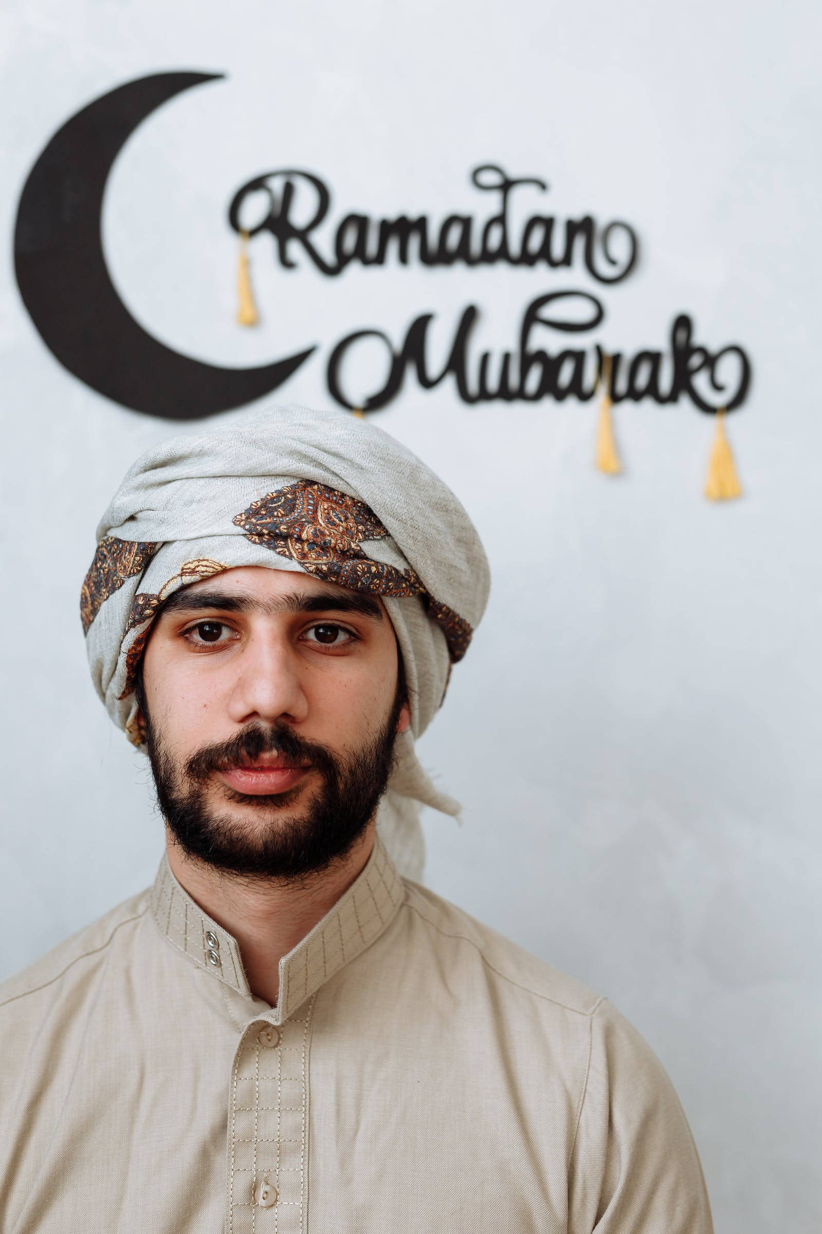 Ramadan Mubarak 桌布 壁紙 — 一名穿著傳統服飾的男子站在帶有金色流蘇的「Ramadan Mubarak」新月標誌前。