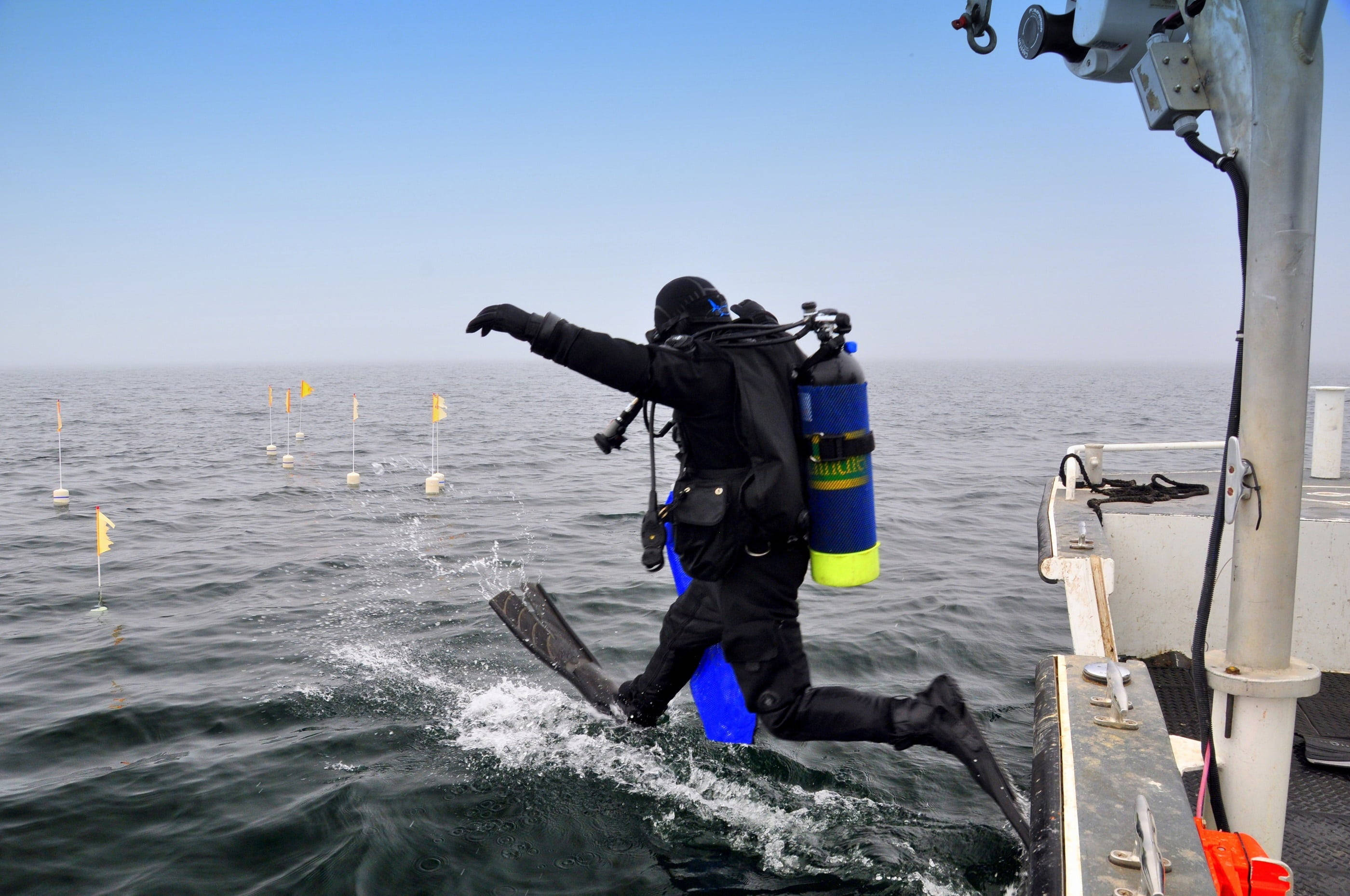 diving 桌布 壁紙 — 一名穿著全套裝備的水肺潛水員從船上跳入海洋，遠處可見黃色浮標。