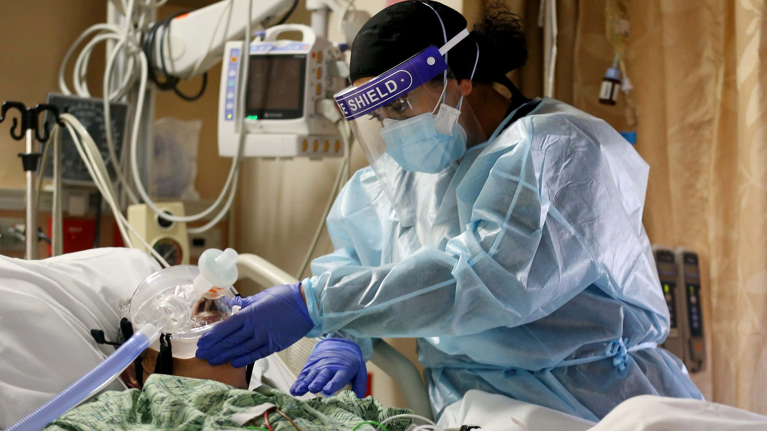 Hospital 桌布 壁紙 — 一名穿著全套防護裝備的醫護人員正在調整醫院病床上病人的呼吸器面罩，周圍是醫療設備。