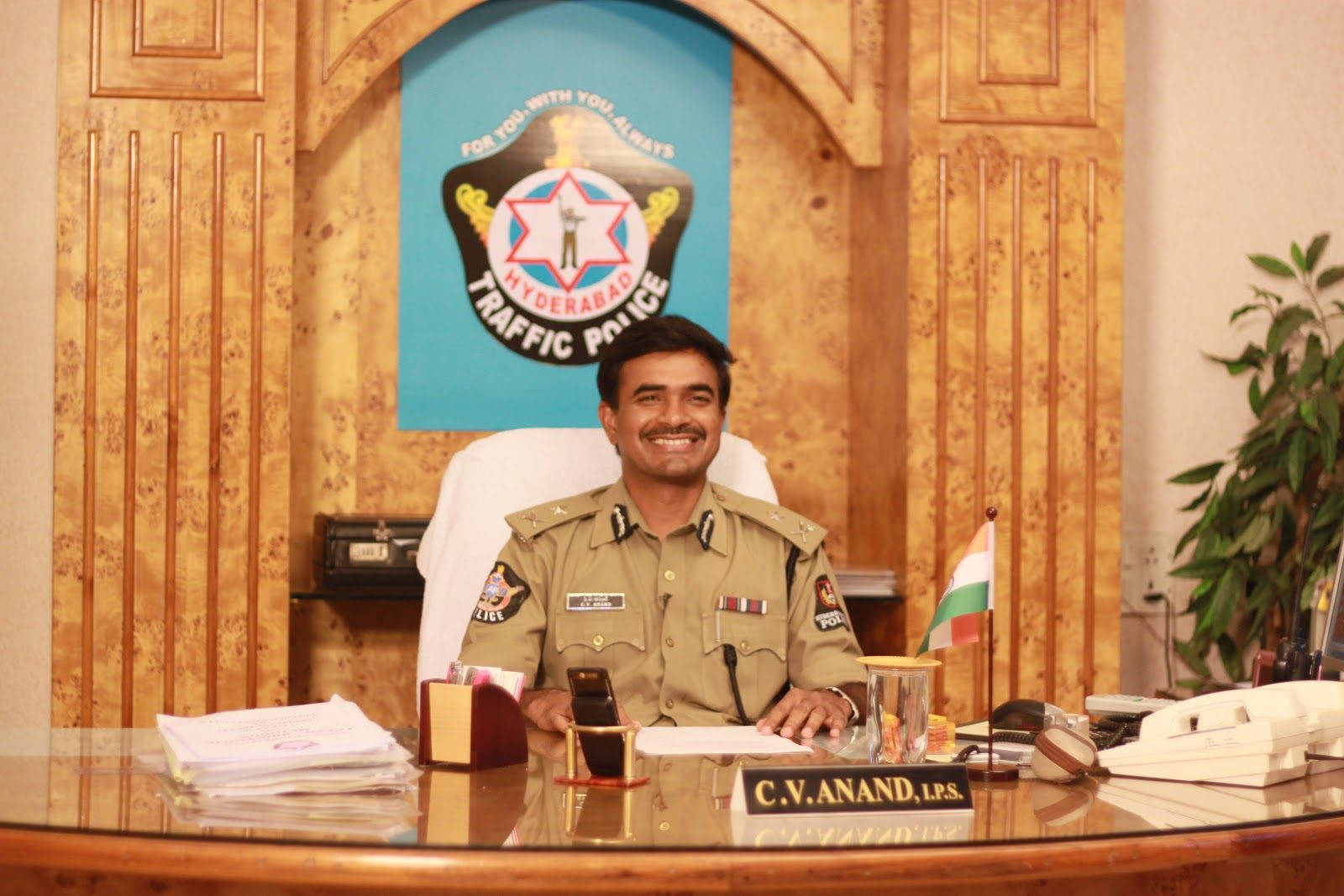 Indian 桌布 壁紙 — 一名穿著制服、面帶微笑的交通警察坐在一張拋光木桌後，身處一間官方辦公室，牆上掛有海德拉巴交通警察標誌。