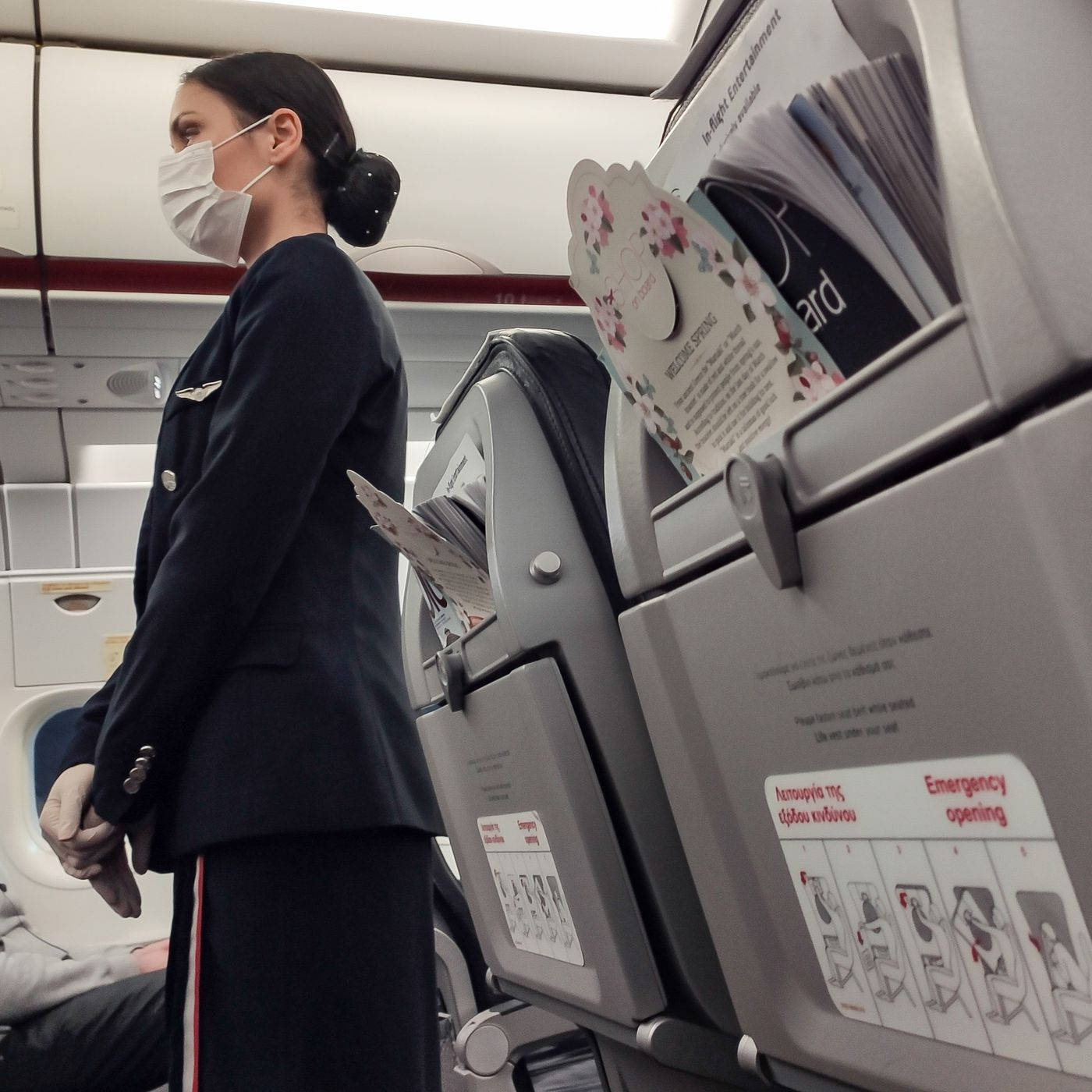 Airport 桌布 壁紙 — 一名穿著制服和口罩的空中服務員站在飛機座位旁，手持雜誌，機艙內有頭頂行李架和安全指示。
