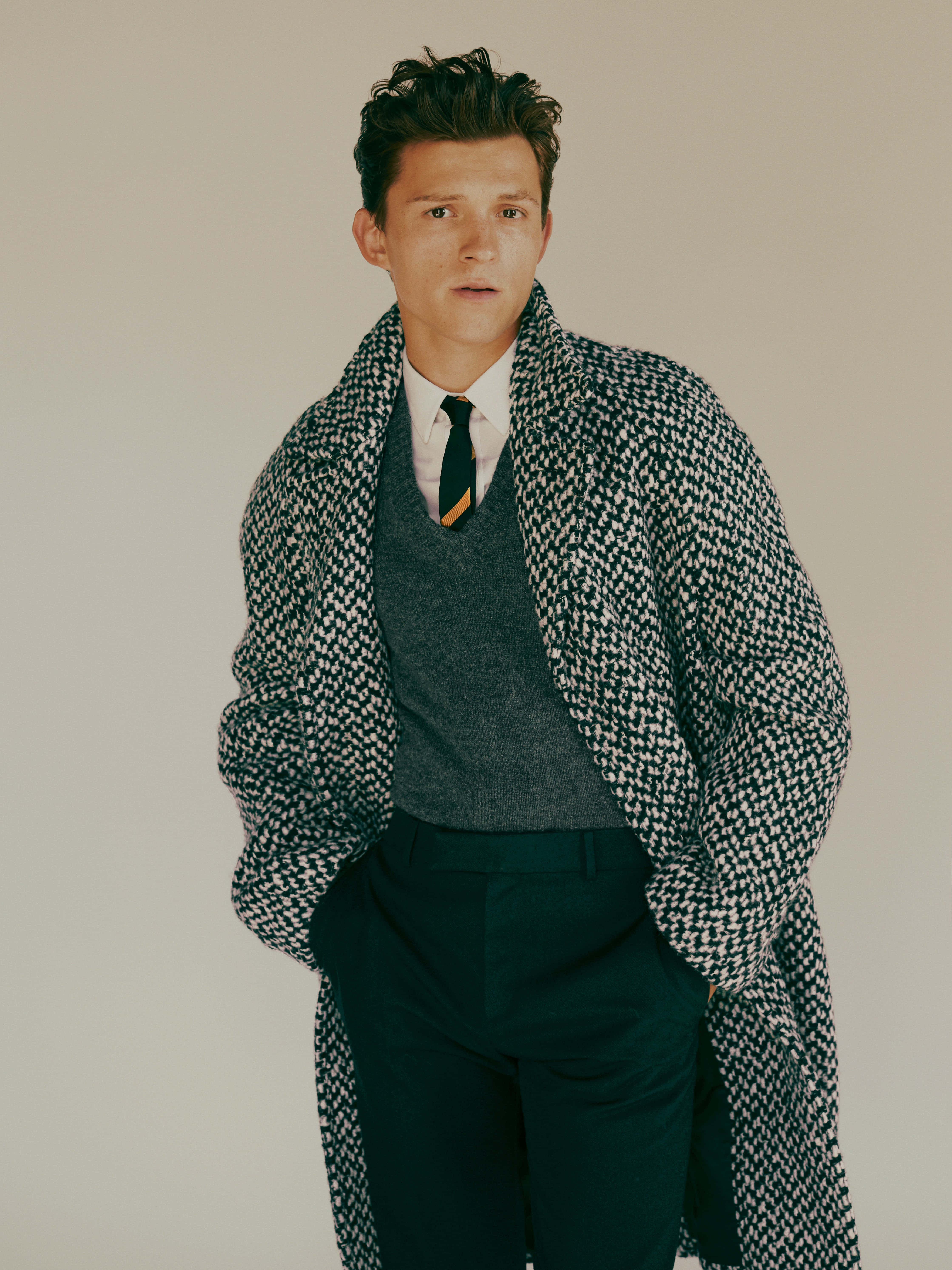 Tom Holland Aesthetic 桌布 壁紙 — 一名穿著千鳥格大衣和毛衣背心的男子，雙手插袋，站在中性背景前。
