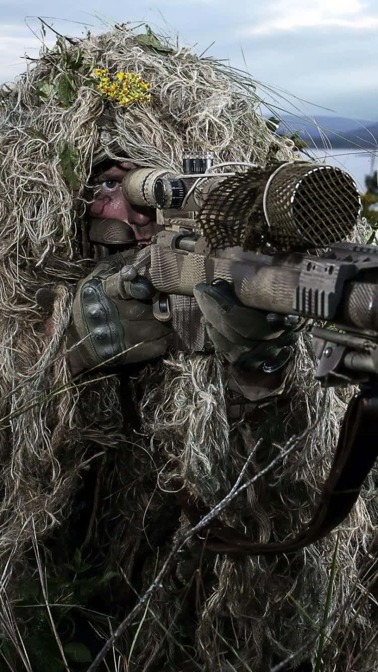 軍事動圖 桌布 壁紙 — 一名穿著吉利服的士兵在草坡上瞄準狙擊步槍，俯瞰遠處的湖泊，天空多雲。