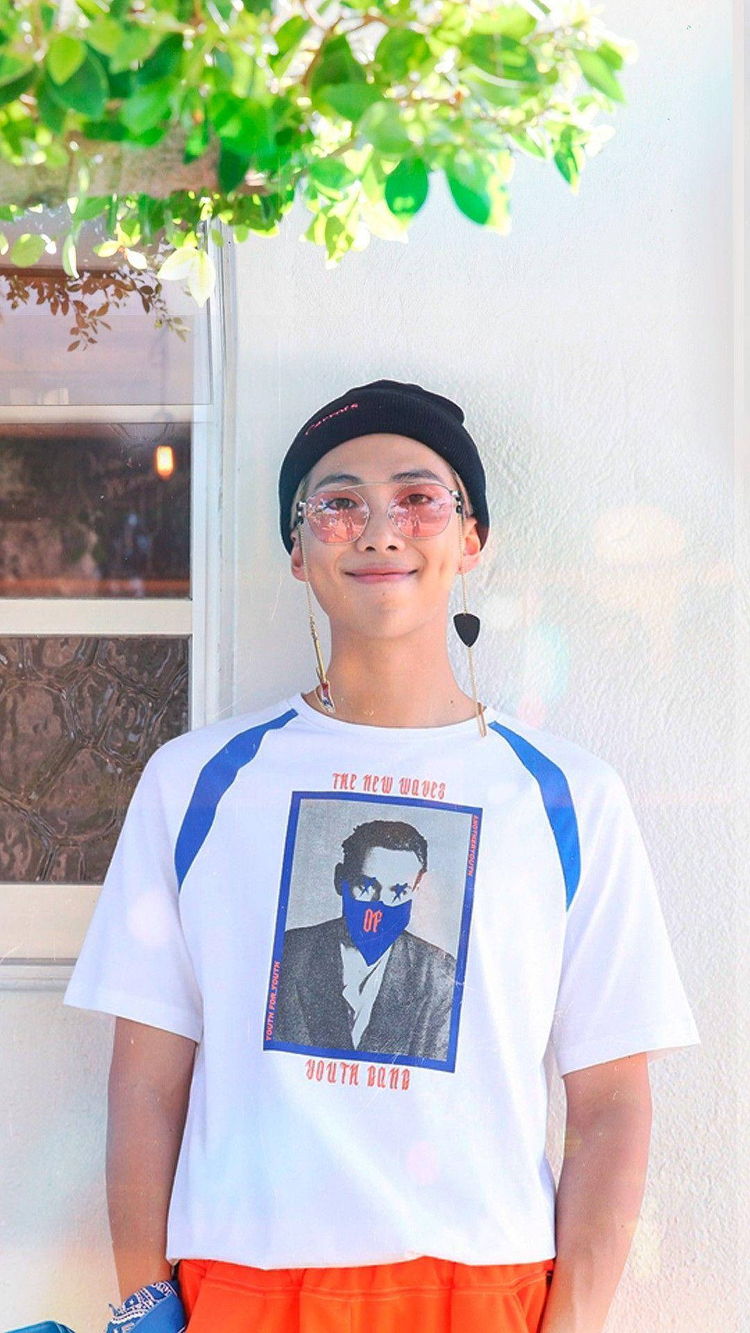 Namjoon 桌布 壁紙 — 一名穿著圖案T恤和毛帽的年輕男子在明亮的白色牆壁前，在翠綠的樹枝下微笑。