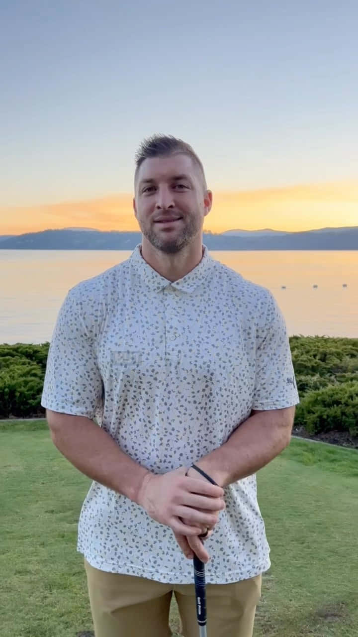 Tim Tebow 桌布 壁紙 — 一名穿著圖案襯衫的男子在日落時分，以平靜的湖泊和遠山為背景，在草地上揮舞高爾夫球桿。