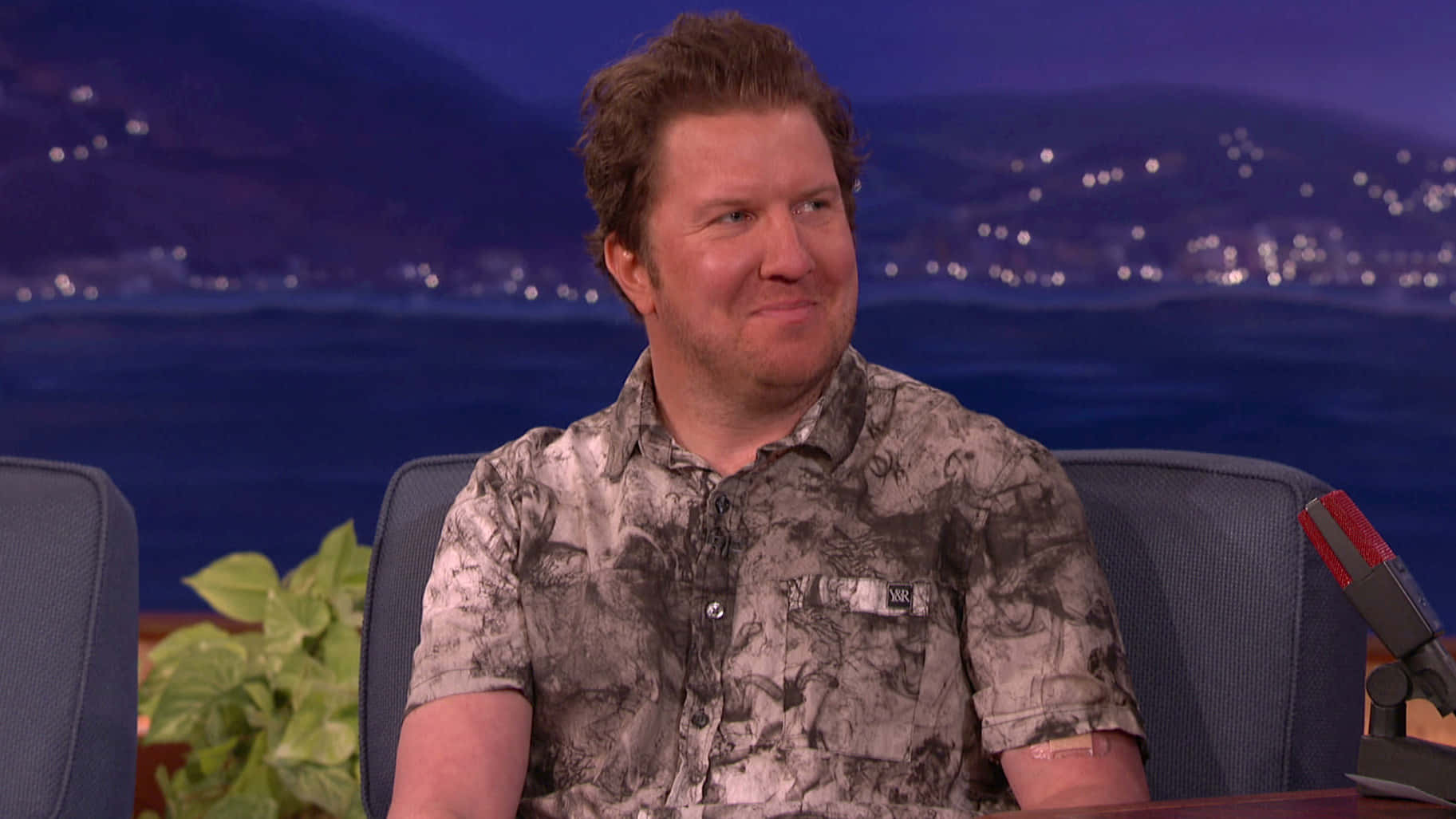 Nick Swardson 桌布 壁紙 — 一名穿著圖案襯衫的男子坐在脫口秀佈景中，背景是夜景城市景觀和水面。