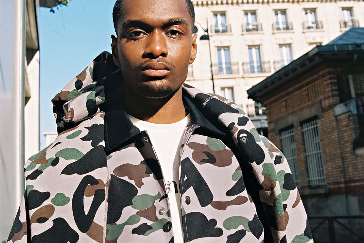 Bape 動態 桌布 壁紙 — 一名穿著大膽迷彩外套的男子自信地站在陽光普照的城市背景下，背景有歷史建築。