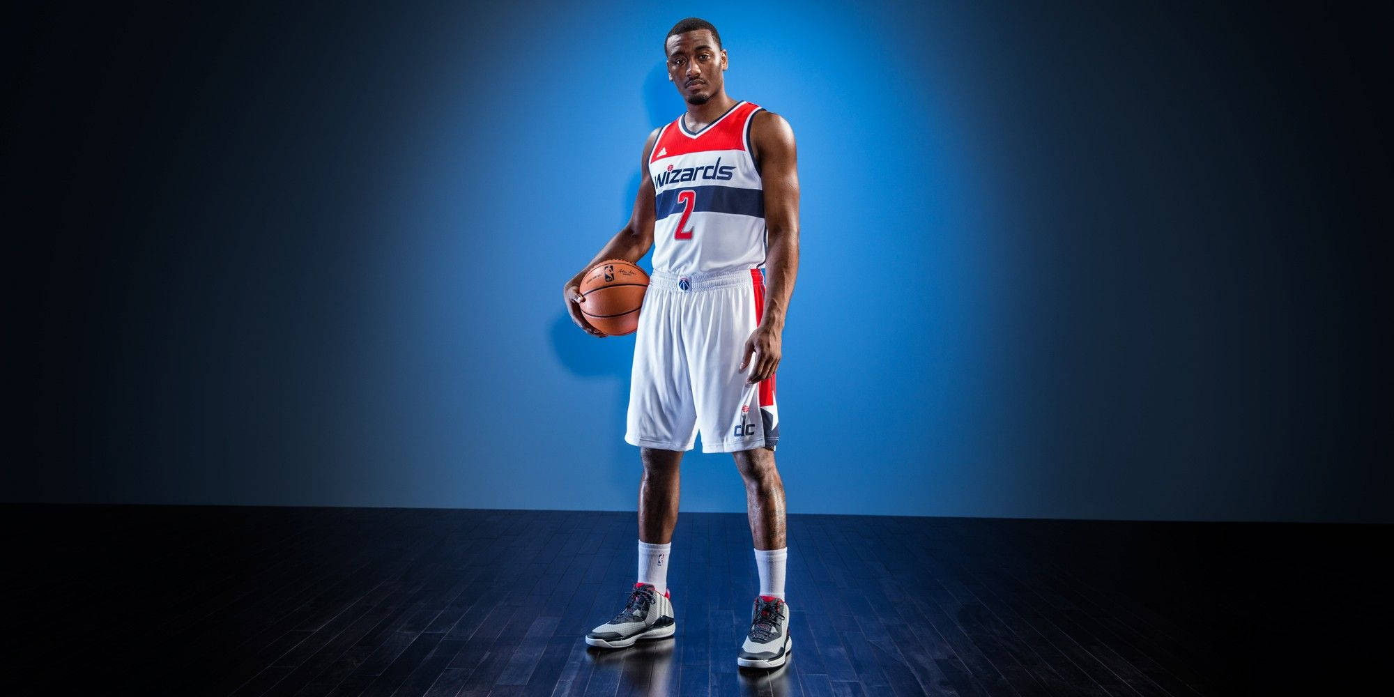 John Wall 桌布 壁紙 — 一名穿著巫師隊球衣的籃球運動員自信地站在漸變藍色攝影棚背景前，手持籃球。