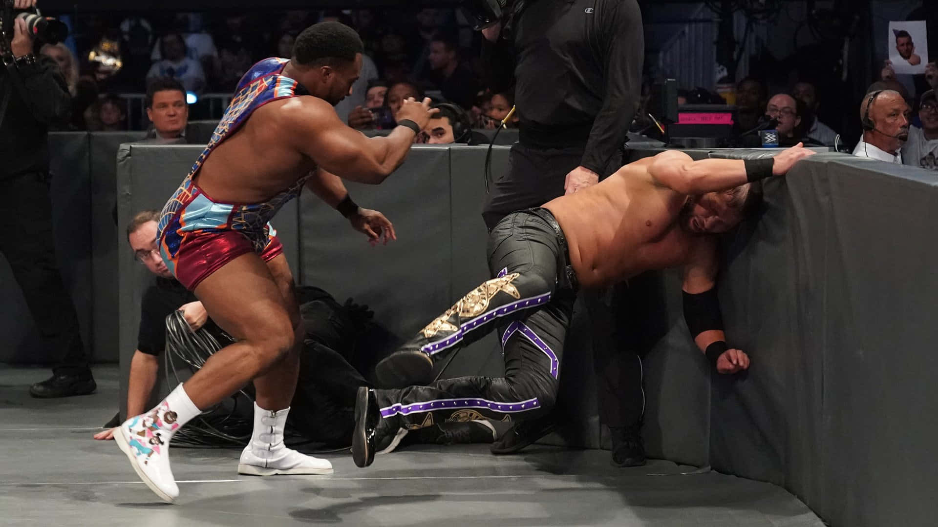 WWE 桌布 壁紙 — 一名穿著彩色短褲的摔角手跳向被壓在擂台邊的對手，裁判在旁仔細觀察。