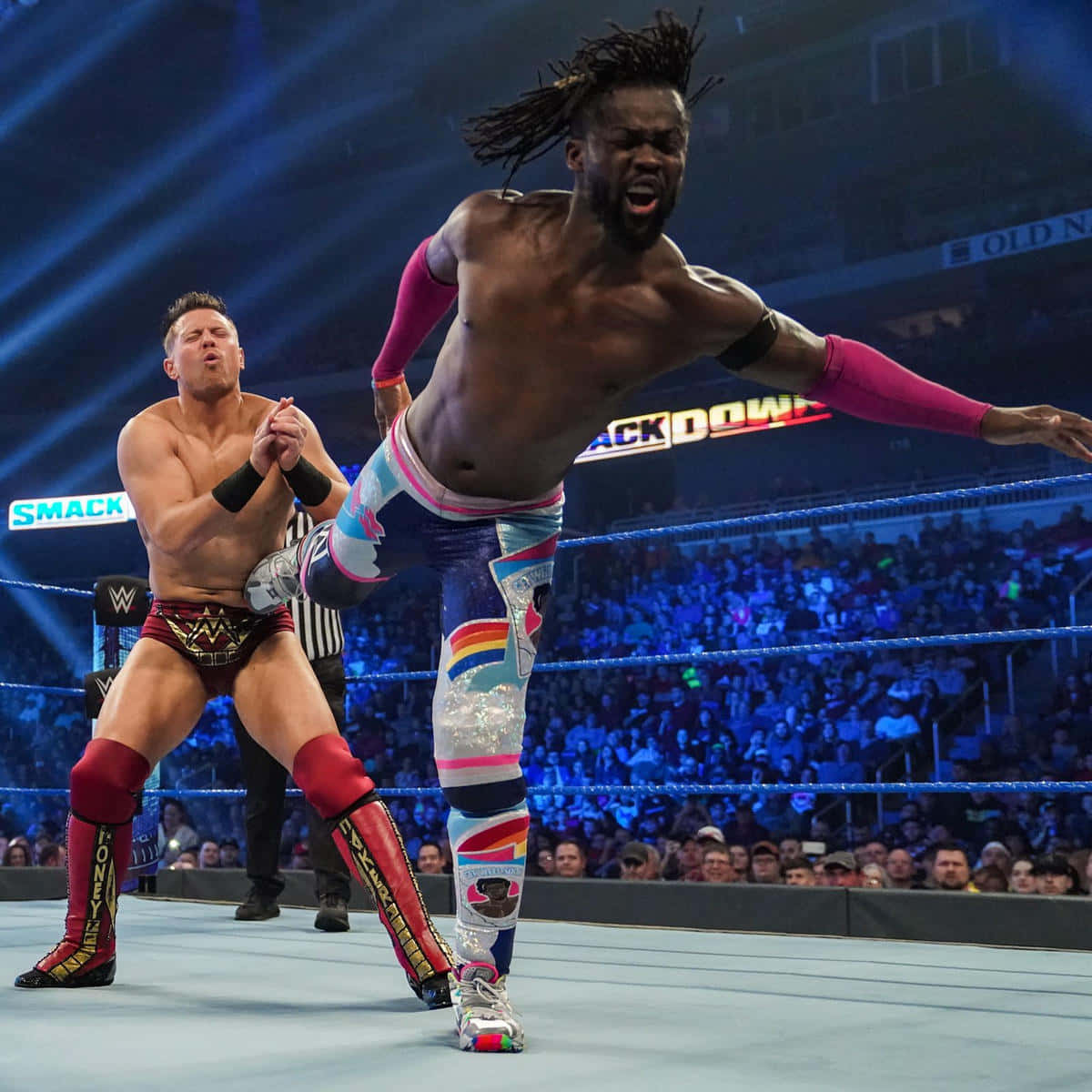 WWE 桌布 壁紙 — 一名穿著彩虹裝備的摔角手在半空中躍起，在燈光明亮的擂台上攻擊對手，四周是咆哮的粉絲。