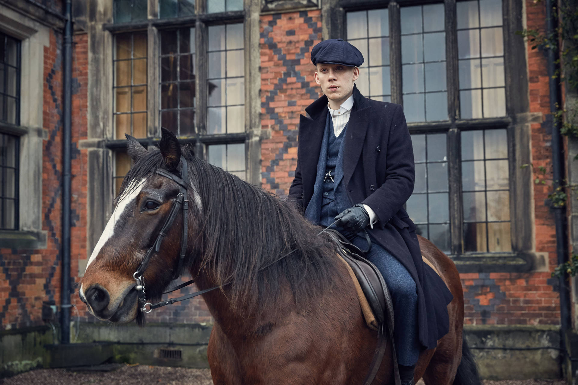 穿著復古外套和帽子的男子，在磚建築附近騎著棕色馬。 — Peaky Blinders 壁紙