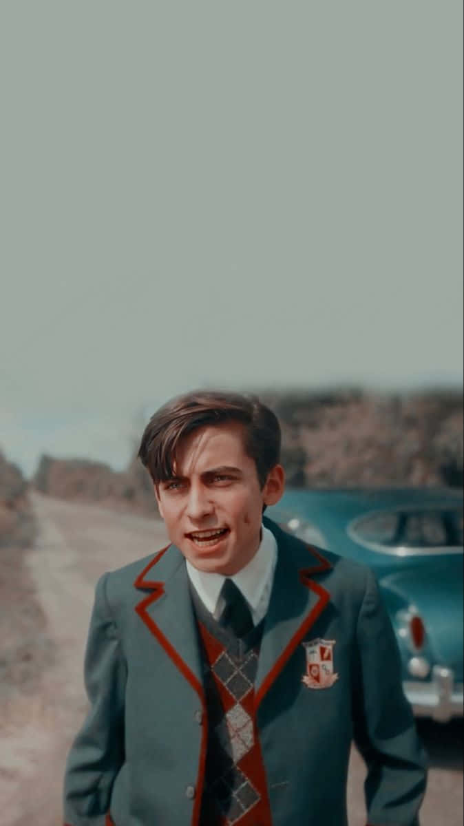Five Umbrella Academy 桌布 壁紙 — 一名穿著復古校服的年輕男子站在灰塵滿佈的道路上，旁邊是一輛青綠色的經典汽車，天空蒼白。