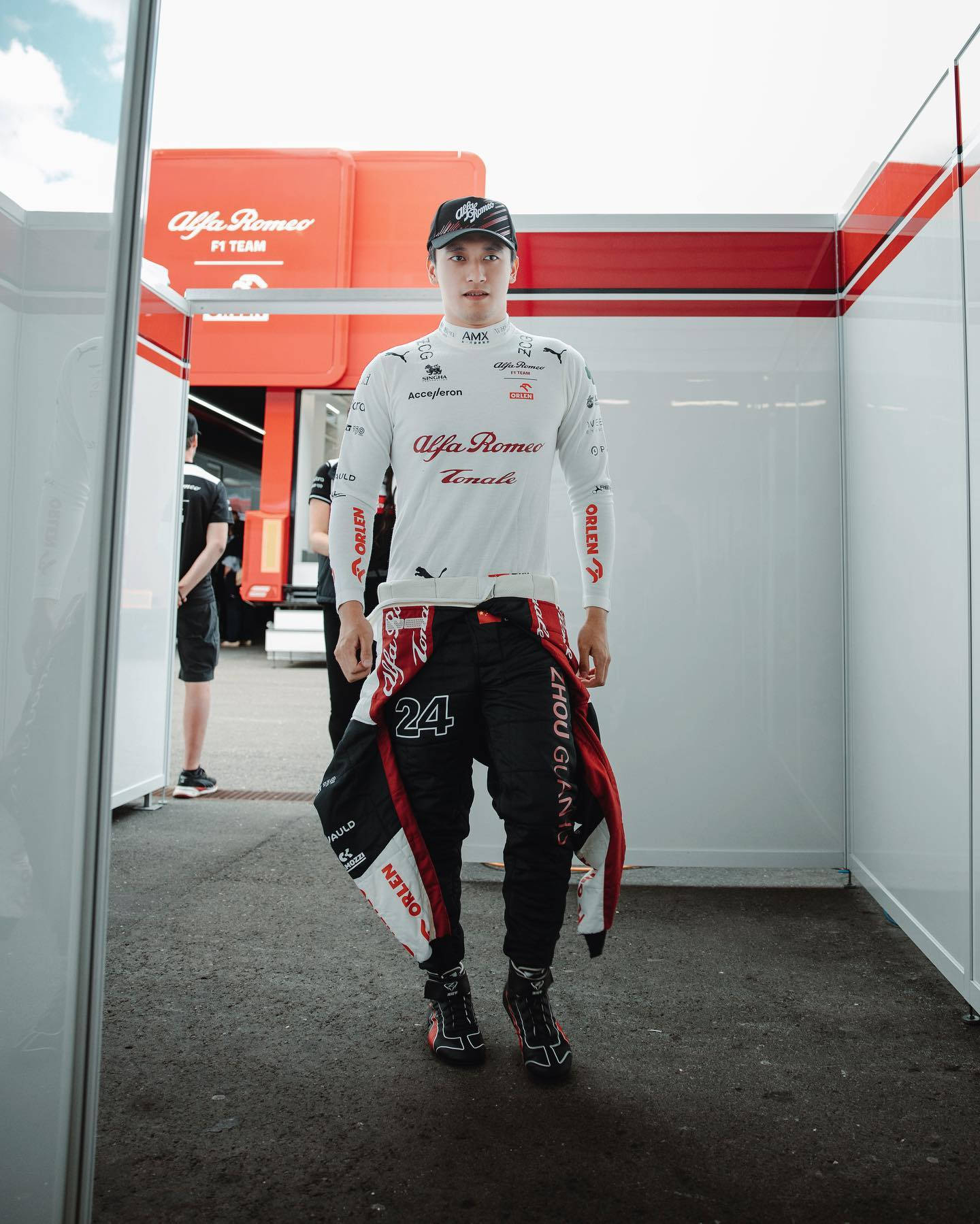 F1 桌布 壁紙 — 一名穿著愛快羅密歐賽車裝備的F1車手自信地走過帶有紅白品牌標識的車隊車庫。