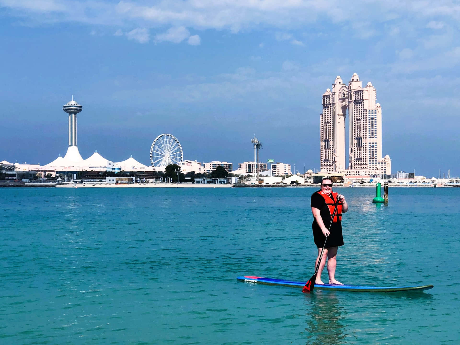 Paddleboarding 桌布 壁紙 — 一名穿著救生衣的人站在槳板上，身處碧綠色的水面，背景是城市天際線，包括摩天輪和高樓大廈，在明亮的藍天下。