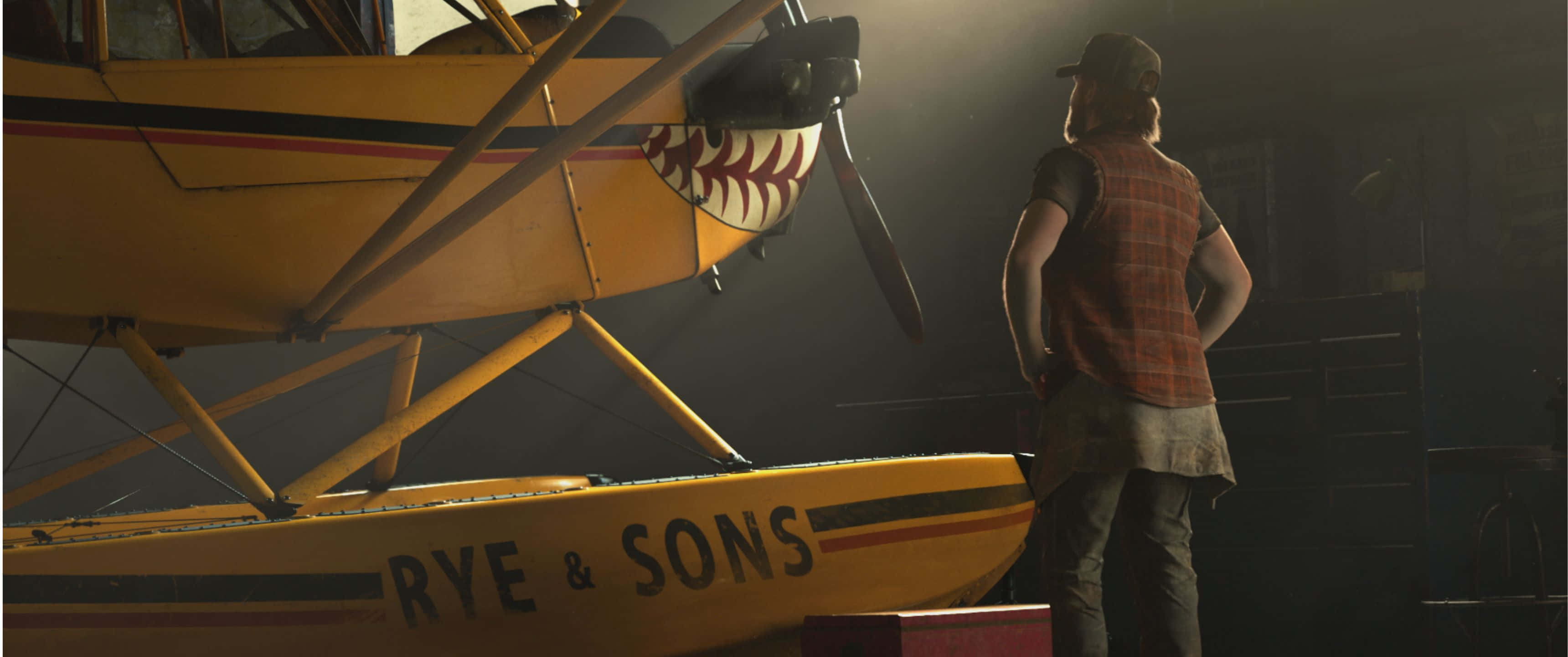 3440x1440p Far Cry 5 桌布 壁紙 — 一名穿著格子背心的男子站在昏暗的機庫裡，旁邊是一架黃色水上飛機，螺旋槳以棒球為主題，機身上寫著「RYE & SONS」。
