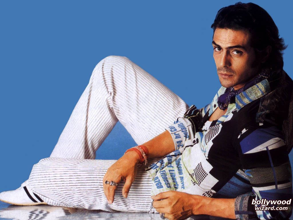 Arjun Rampal 桌布 壁紙 — 一名穿著條紋褲和圖案襯衫的男子，隨意地靠著純藍色背景，直視前方。